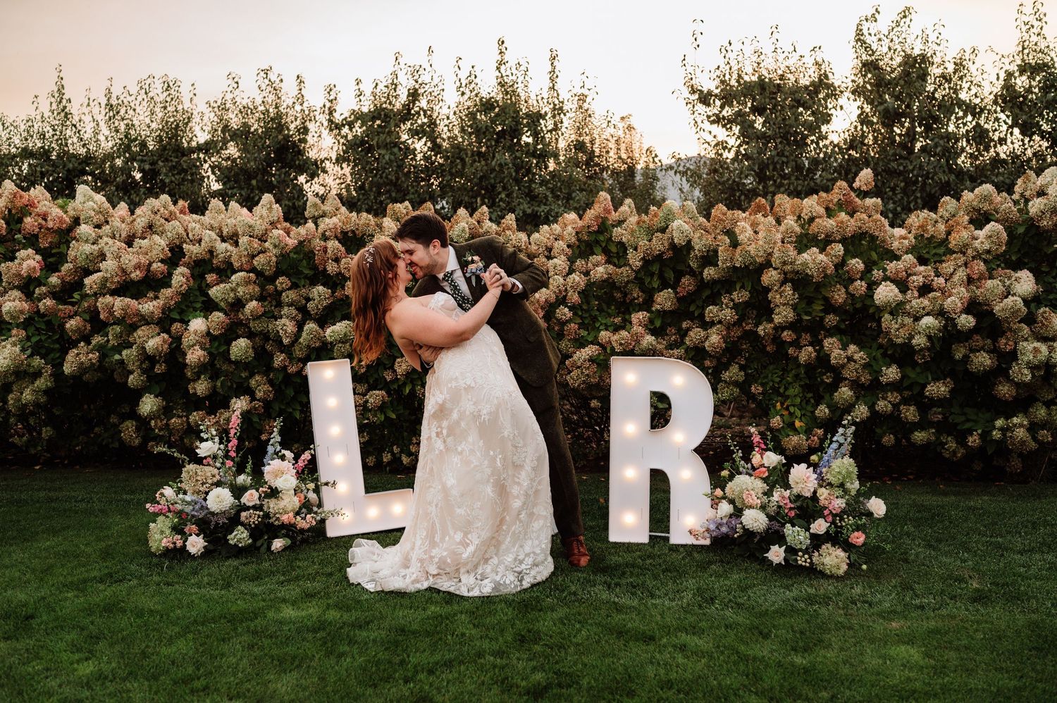 Newlyweds kiss between illuminated L and R marquee letters in front of lush hydrangea backdrop.
