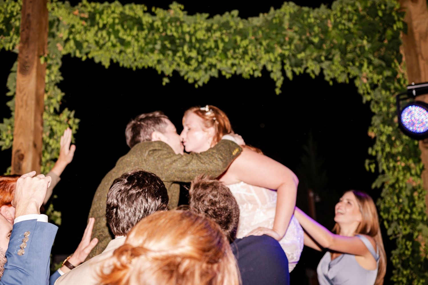 A groom lifts and kisses his bride while surrounded by cheering wedding guests under a vine-covered arch.