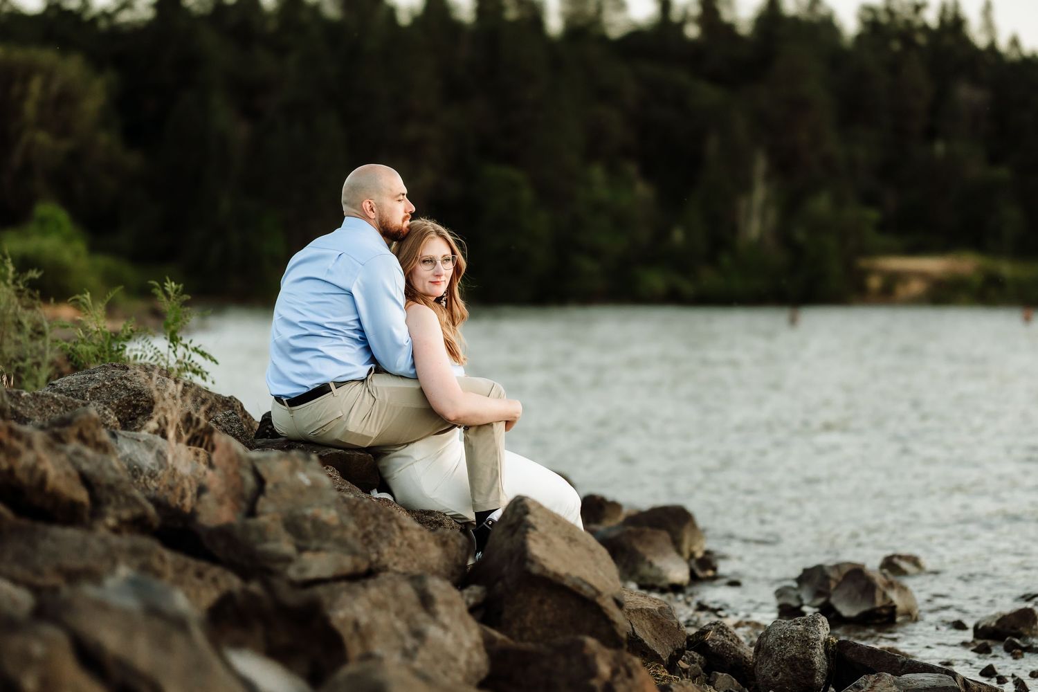 Couple sits together on rocky lakeshore, woman leaning against man in blue shirt at sunset.
