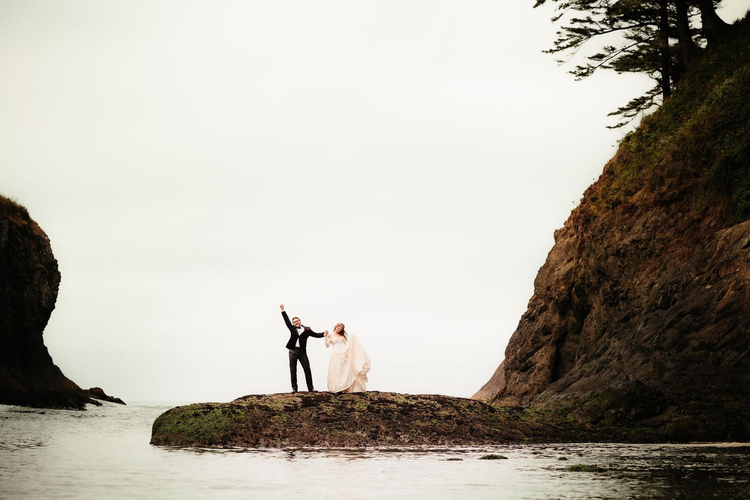 Bride and groom celebrate on coastal rocks surrounded by dramatic ocean cliffs.