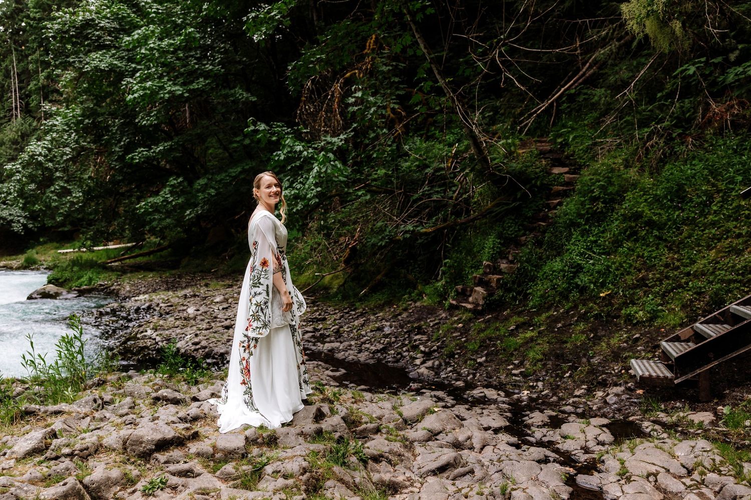 Bride in floral embroidered white gown stands along a rocky riverbank surrounded by lush green forest.