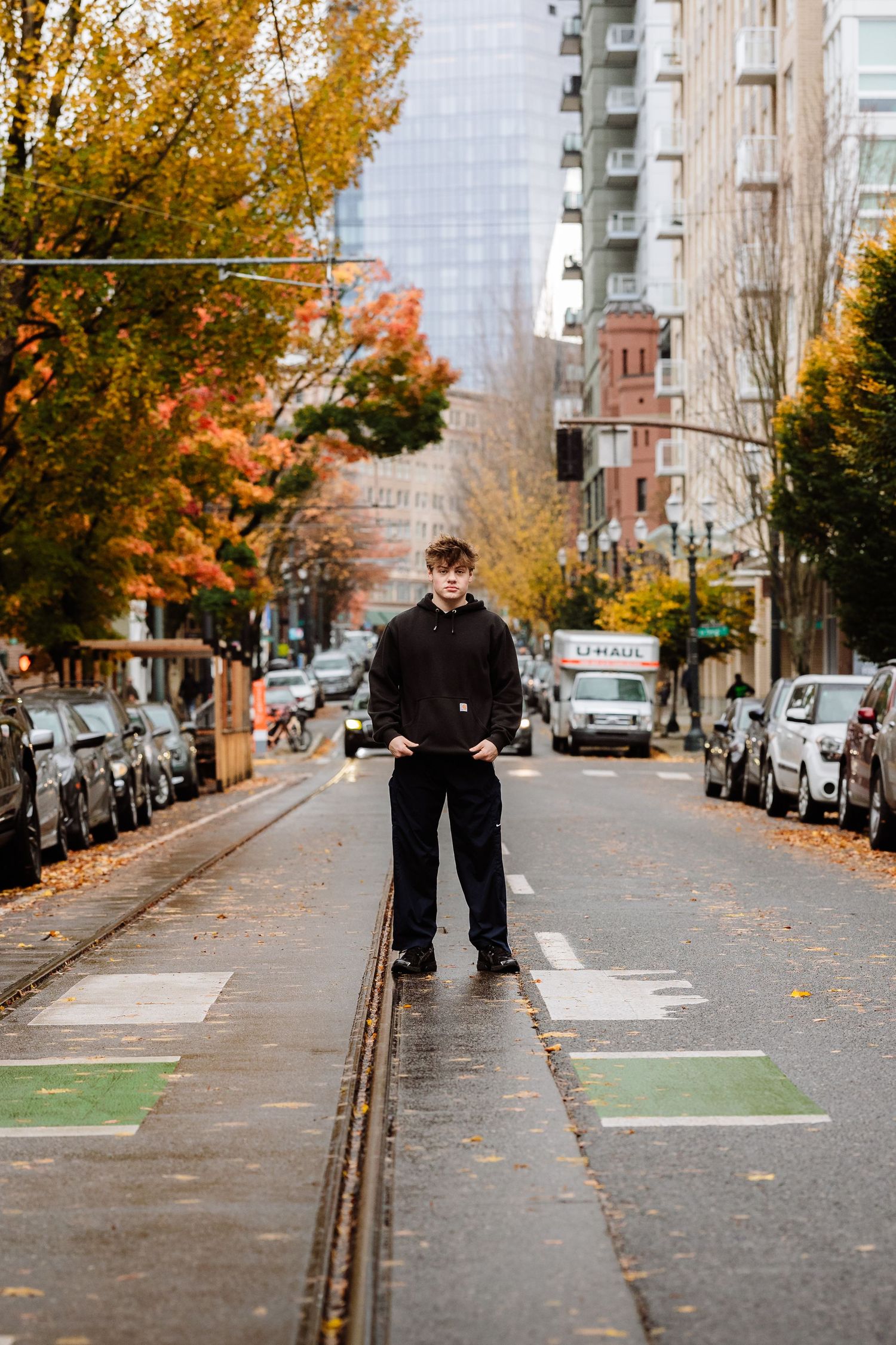 Young man in black outfit standing on urban street railway tracks surrounded by autumn foliage and city buildings.