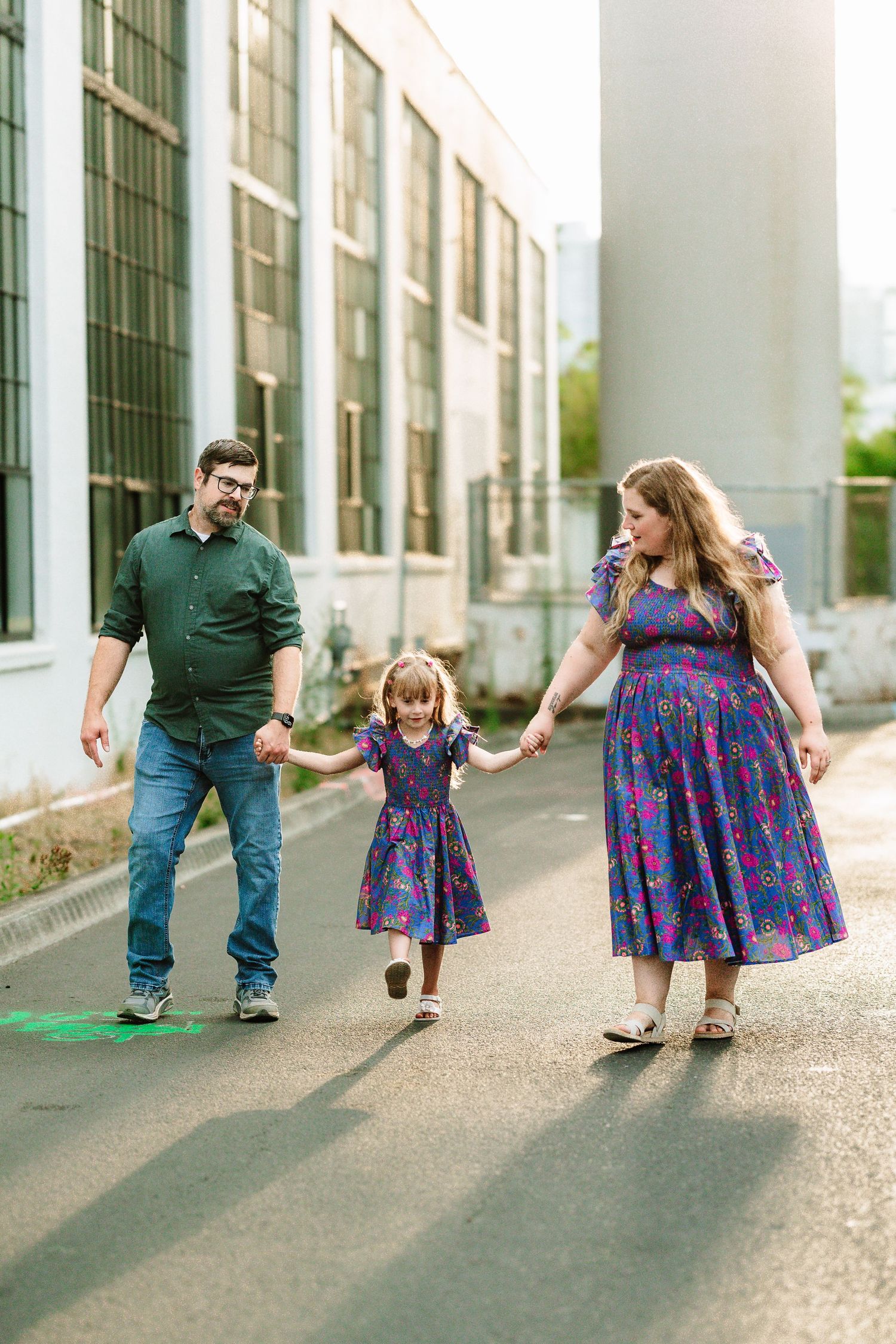 A family of three walks hand-in-hand outdoors, with parents and young girl in matching purple floral dresses near an industrial building.