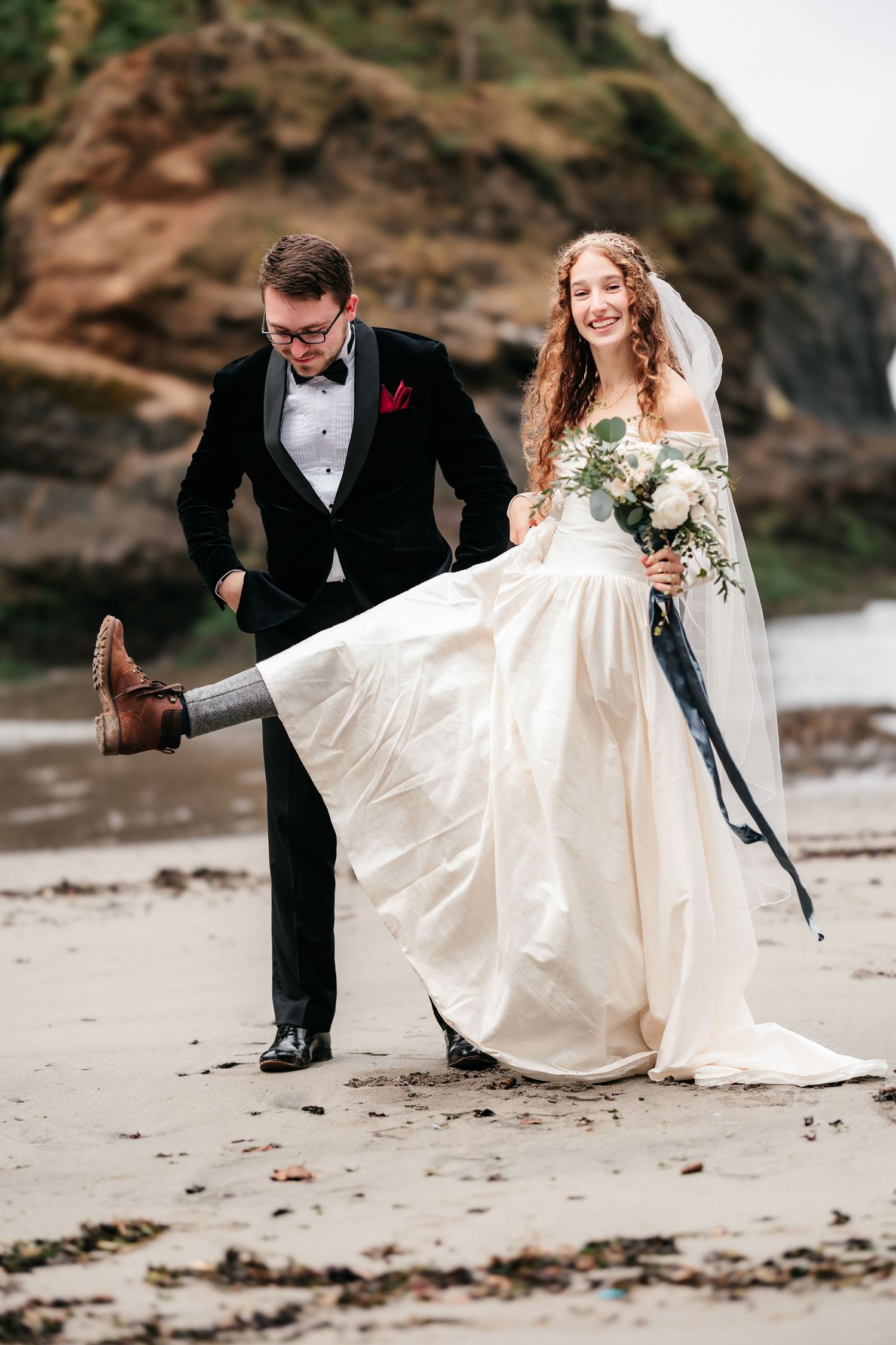 Playful bride and groom laugh together on sandy beach during wedding photos.