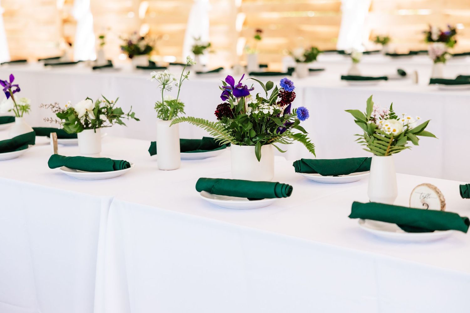 Elegant wedding reception table with white linens, green napkins, and small floral centerpieces in white vases.