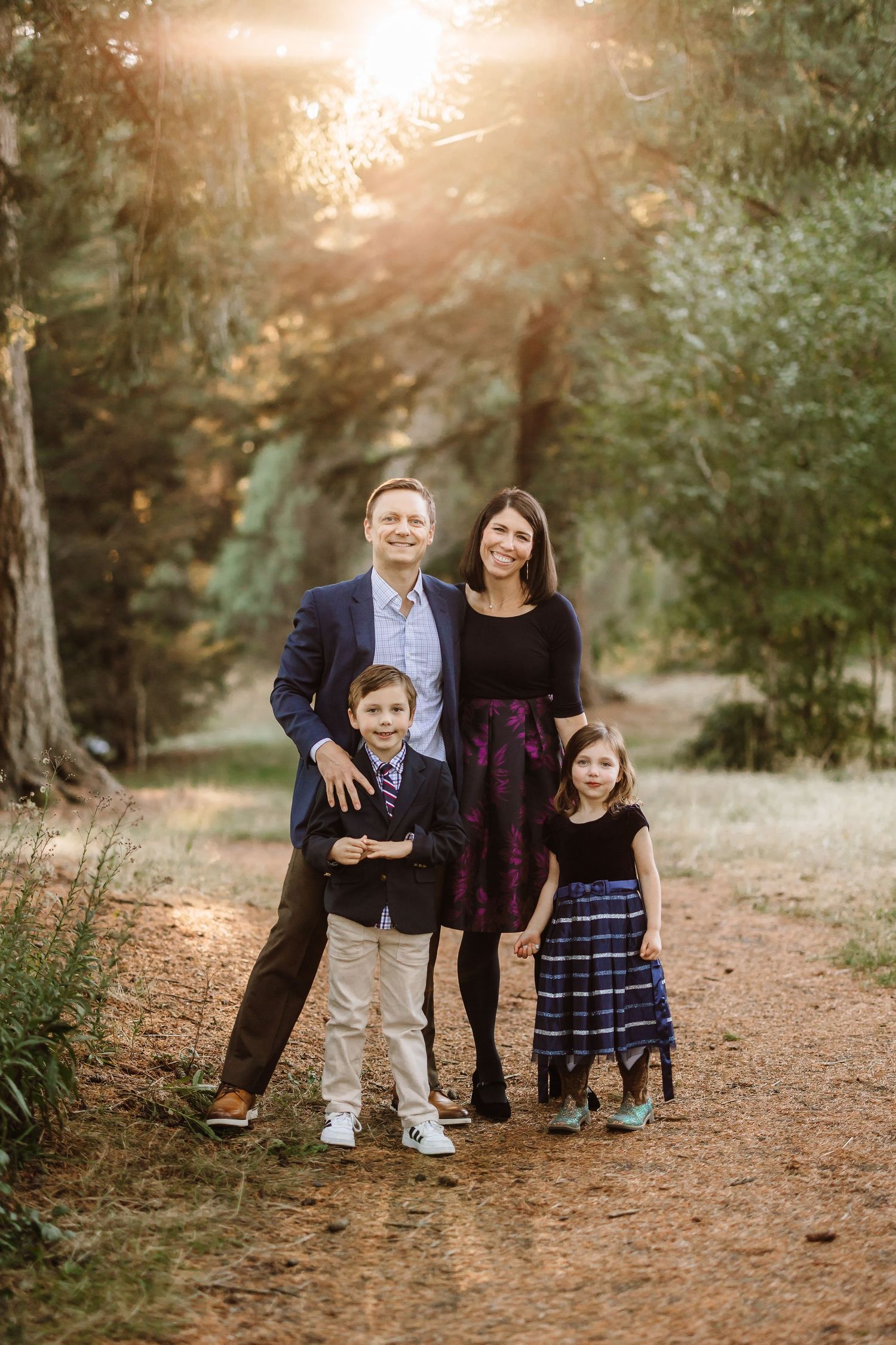 Family of four posing on a sunlit forest path, parents and two children dressed in semi-formal attire for an outdoor portrait.