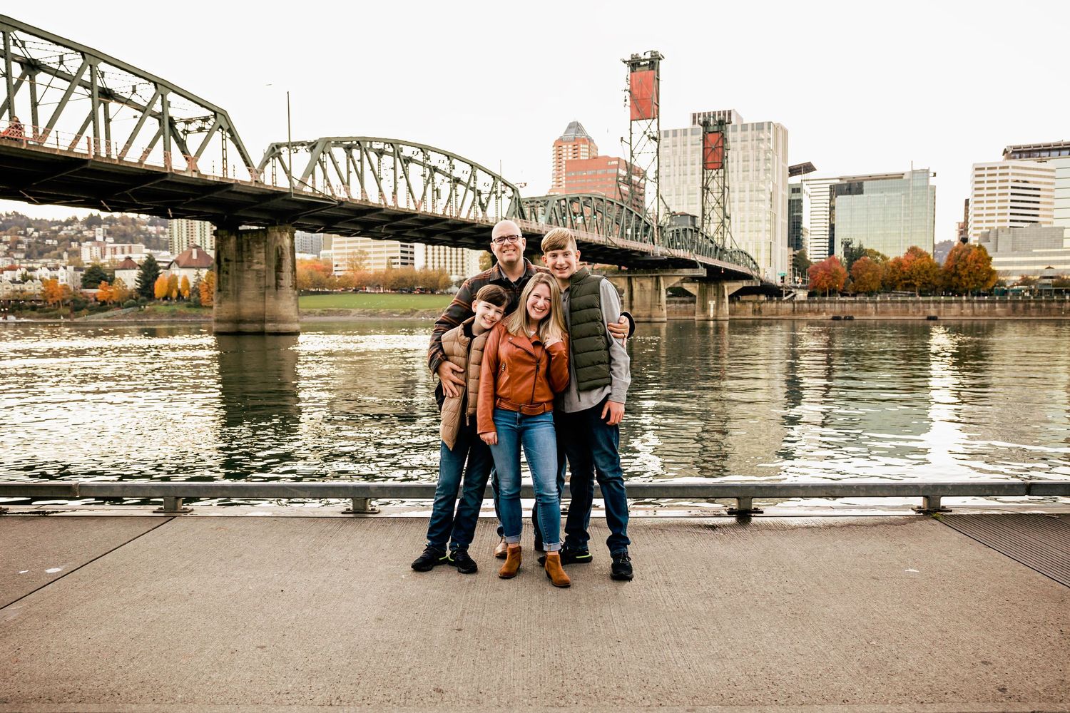 Family of four smiling together on a riverside walkway with a steel bridge and city skyline in the background.