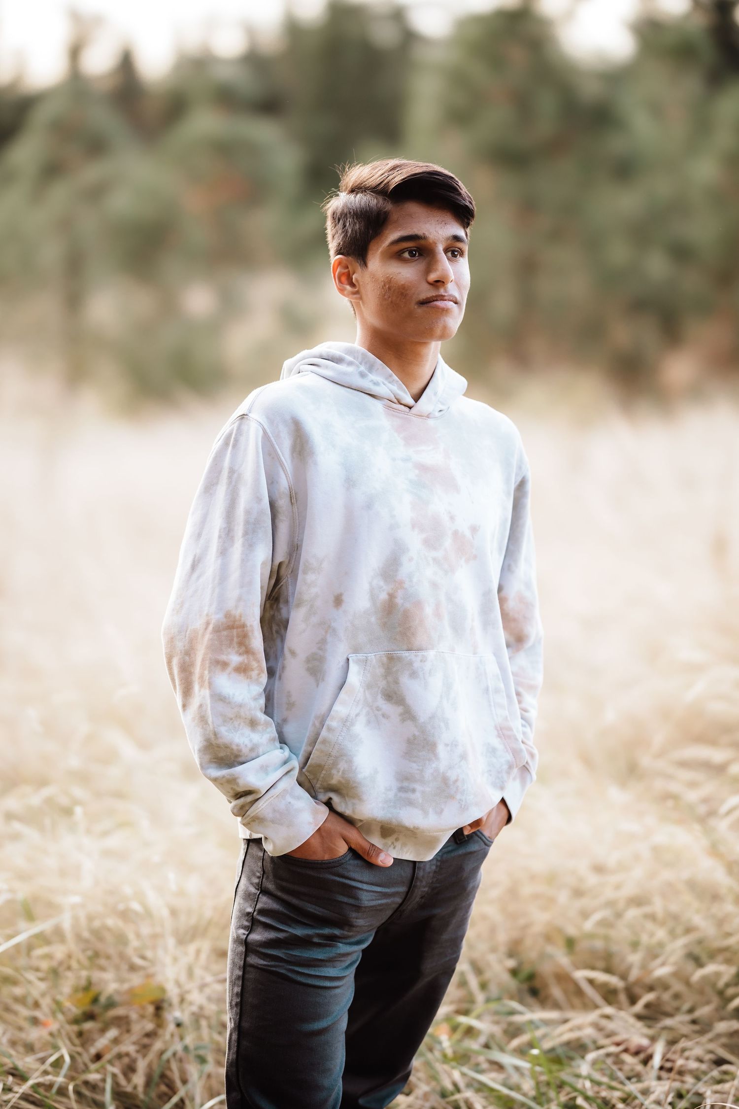 Teen boy in tie-dye hoodie and dark jeans stands in a golden field with evergreen trees in background.
