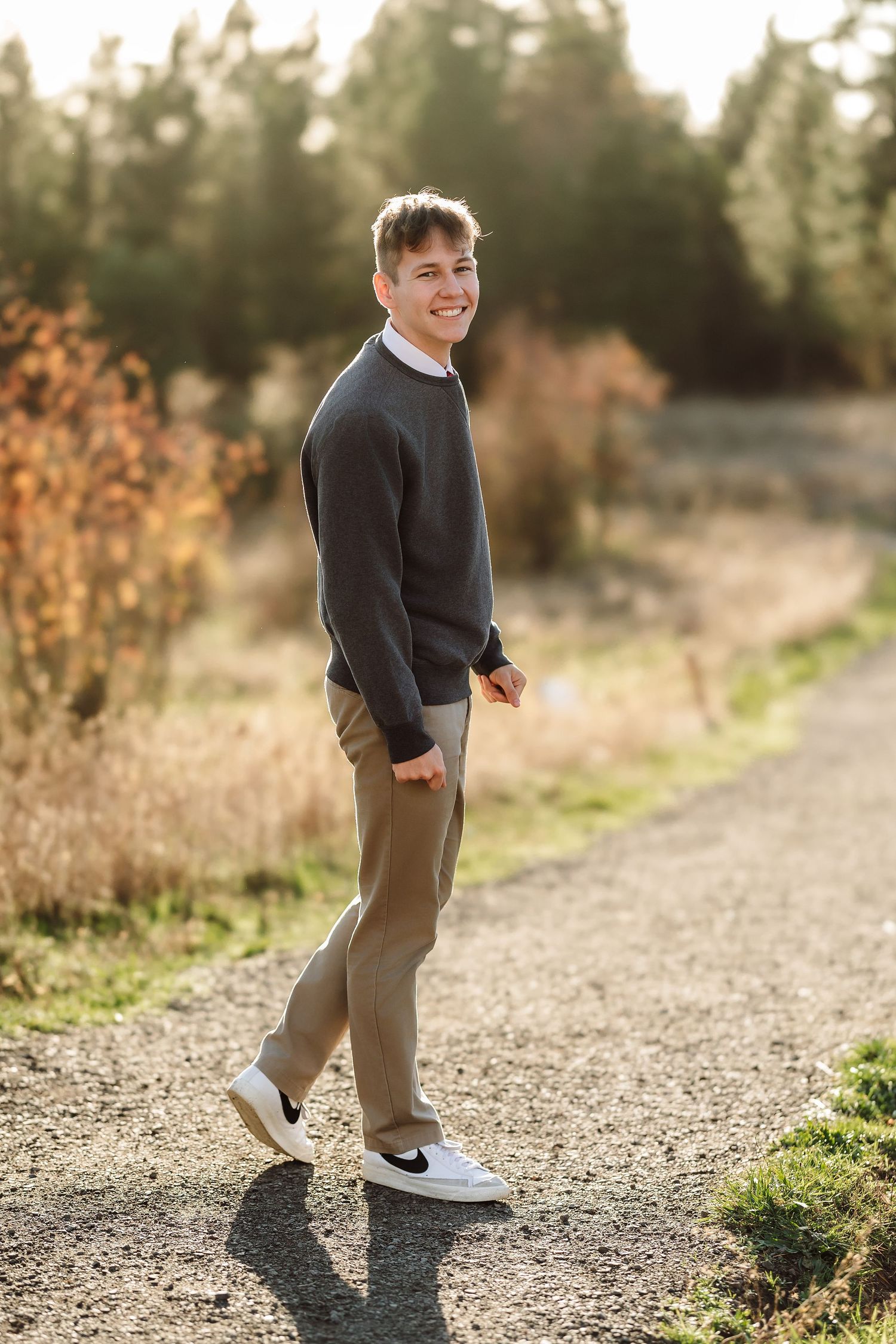 Young man smiling in dark sweater and khaki pants walking on gravel path with golden autumn trees in background.