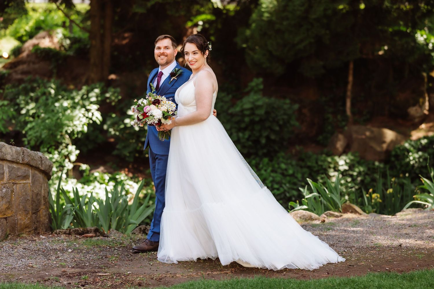 Bride in white gown and groom in blue suit pose together in a sunlit garden with flowers.
