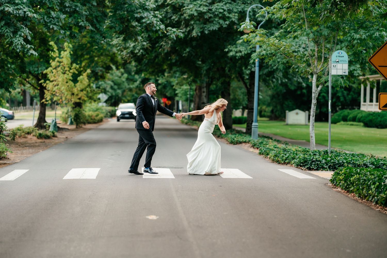 Bride and groom crossing a street on a crosswalk, holding hands and dancing in wedding attire outdoors.