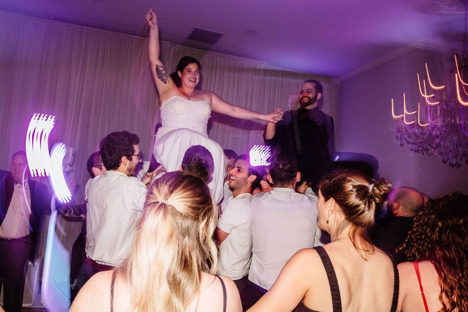Bride lifted by groomsmen during lively hora dance at wedding reception under purple lights.