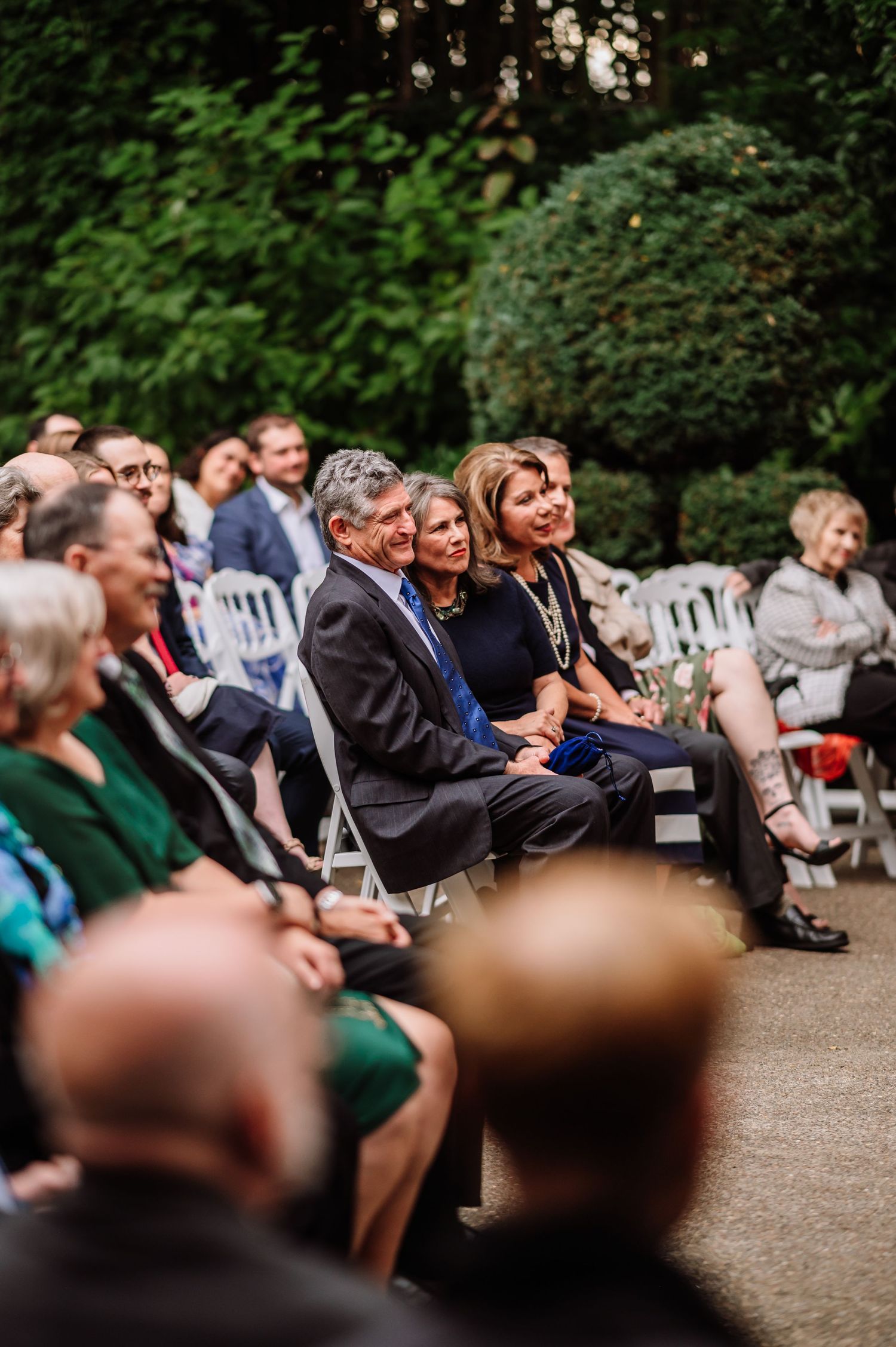 Wedding guests seated in white chairs smile and watch outdoor garden ceremony.