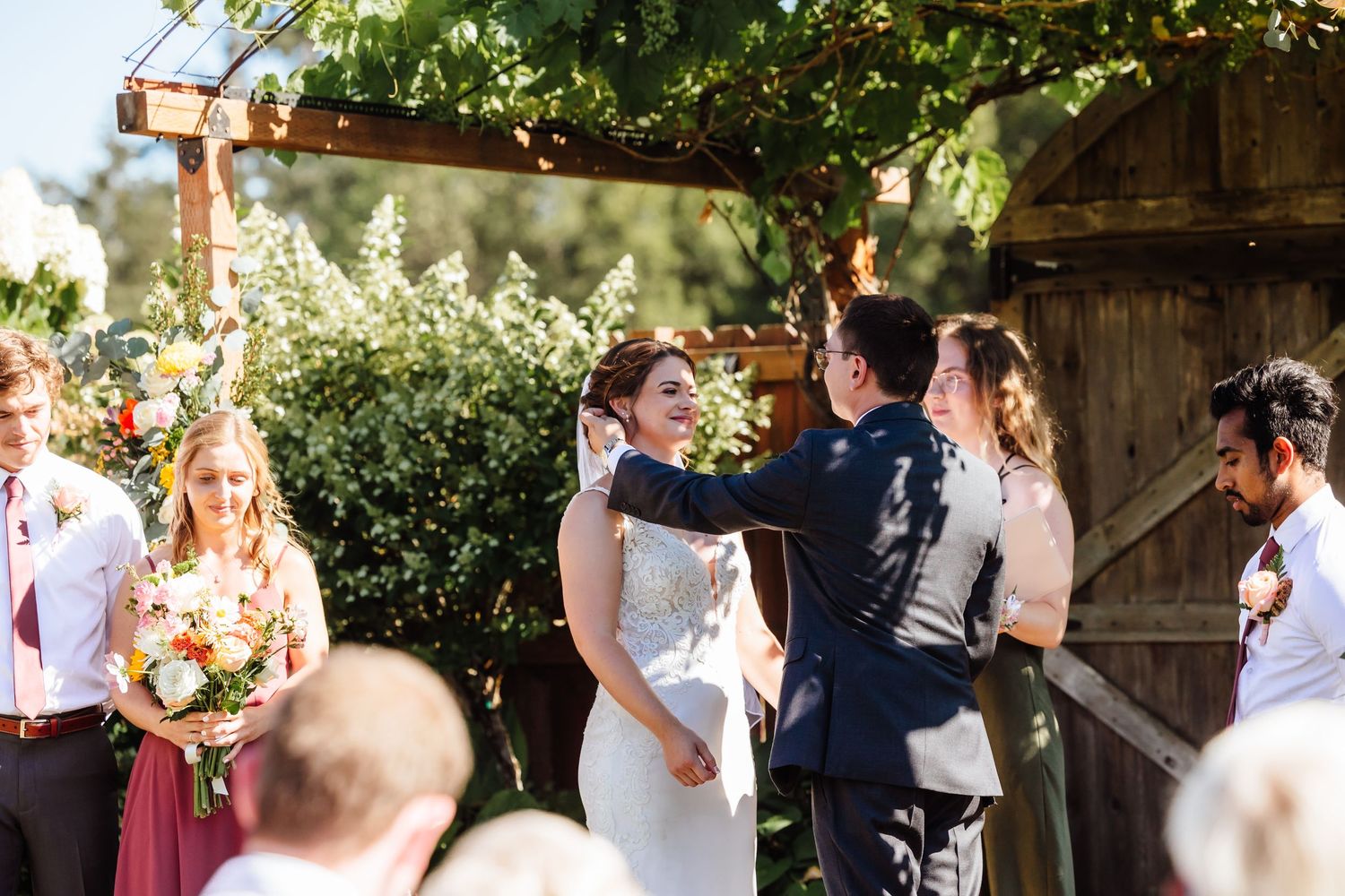 Bride and groom exchange vows under wooden pergola at rustic outdoor wedding ceremony with guests.
