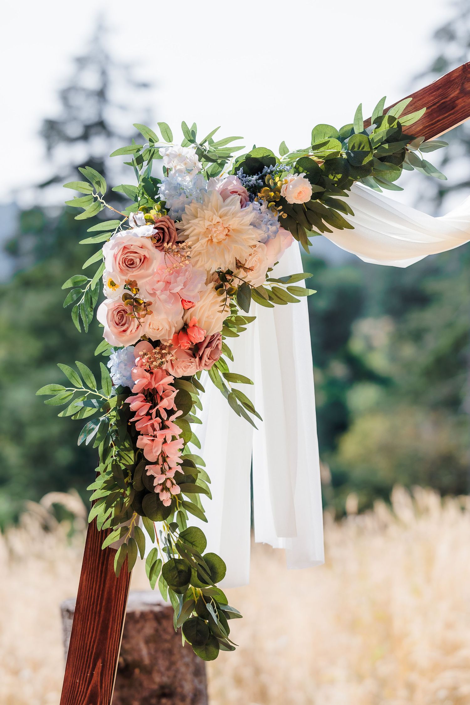 Elegant wedding arch decorated with pink roses, dahlias, and greenery with flowing white fabric outdoors.