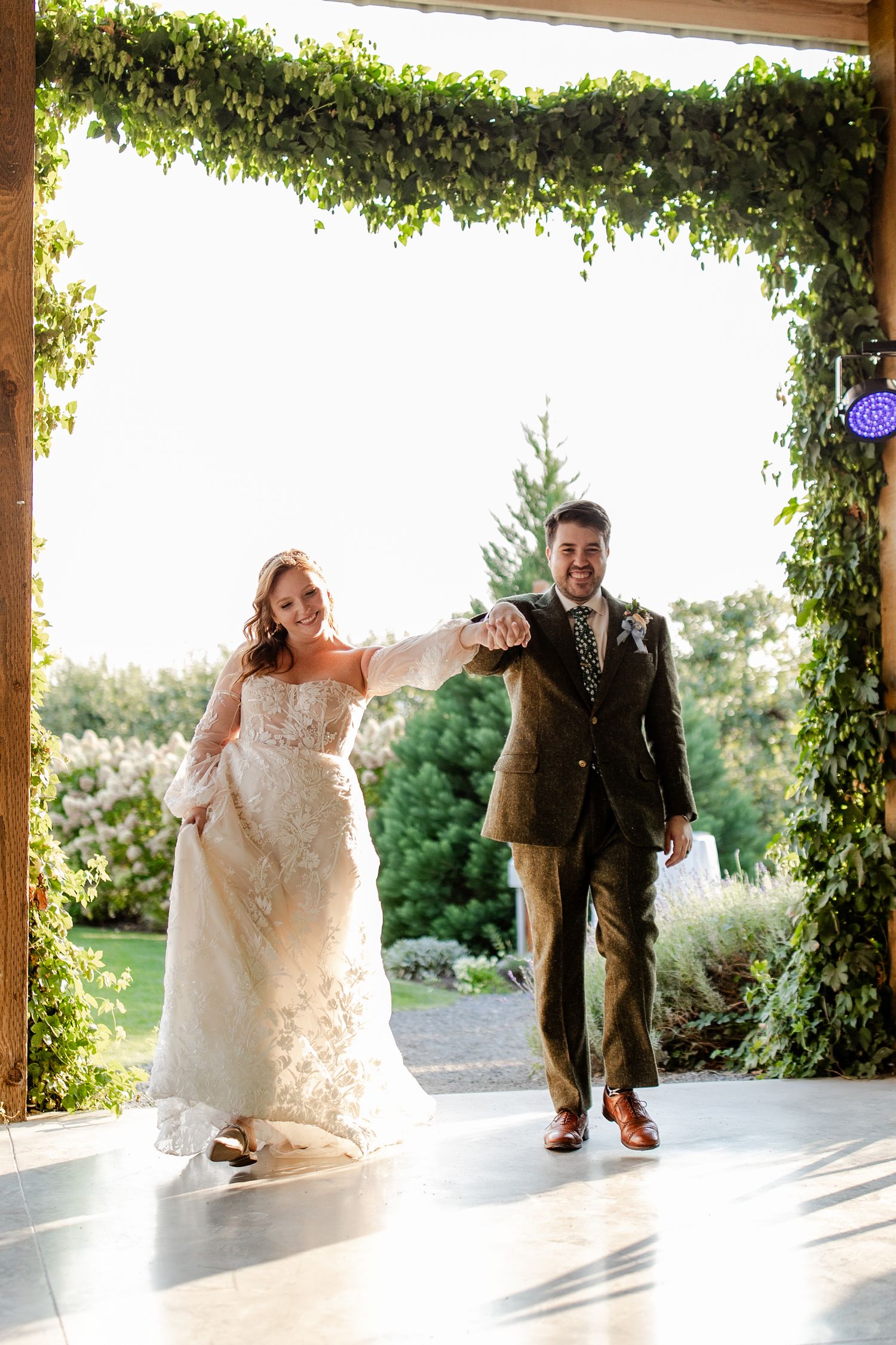 A bride and groom hold hands as they make their grand entrance under a lush ivy-covered archway.