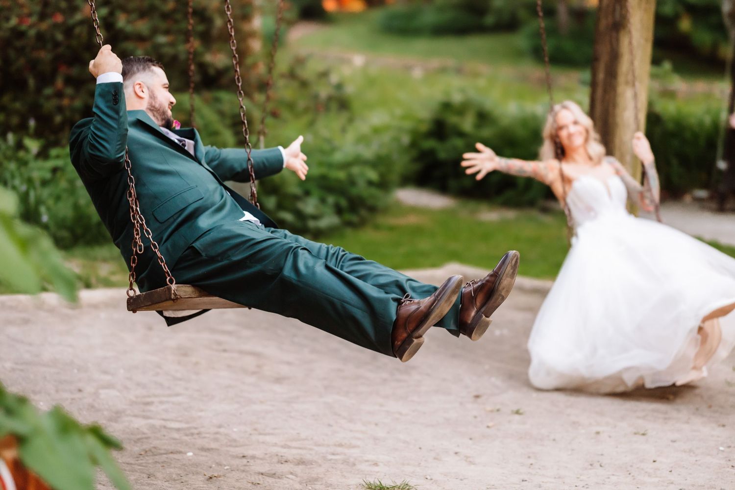 Groom swings on chain swing while bride in white dress rushes toward him in outdoor garden.