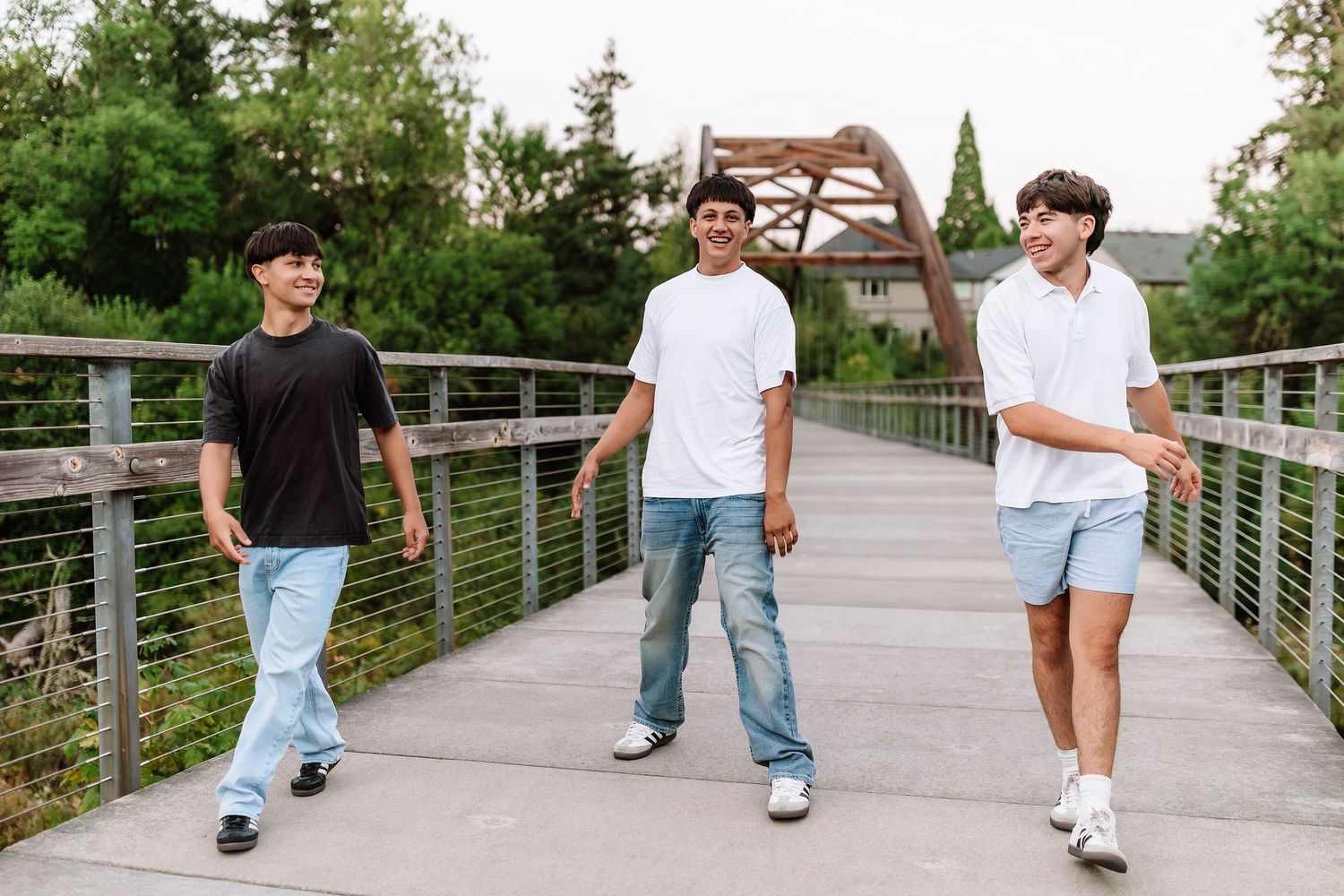 Three teenage boys walking on a wooden bridge outdoors, wearing casual streetwear in black, white, and blue.