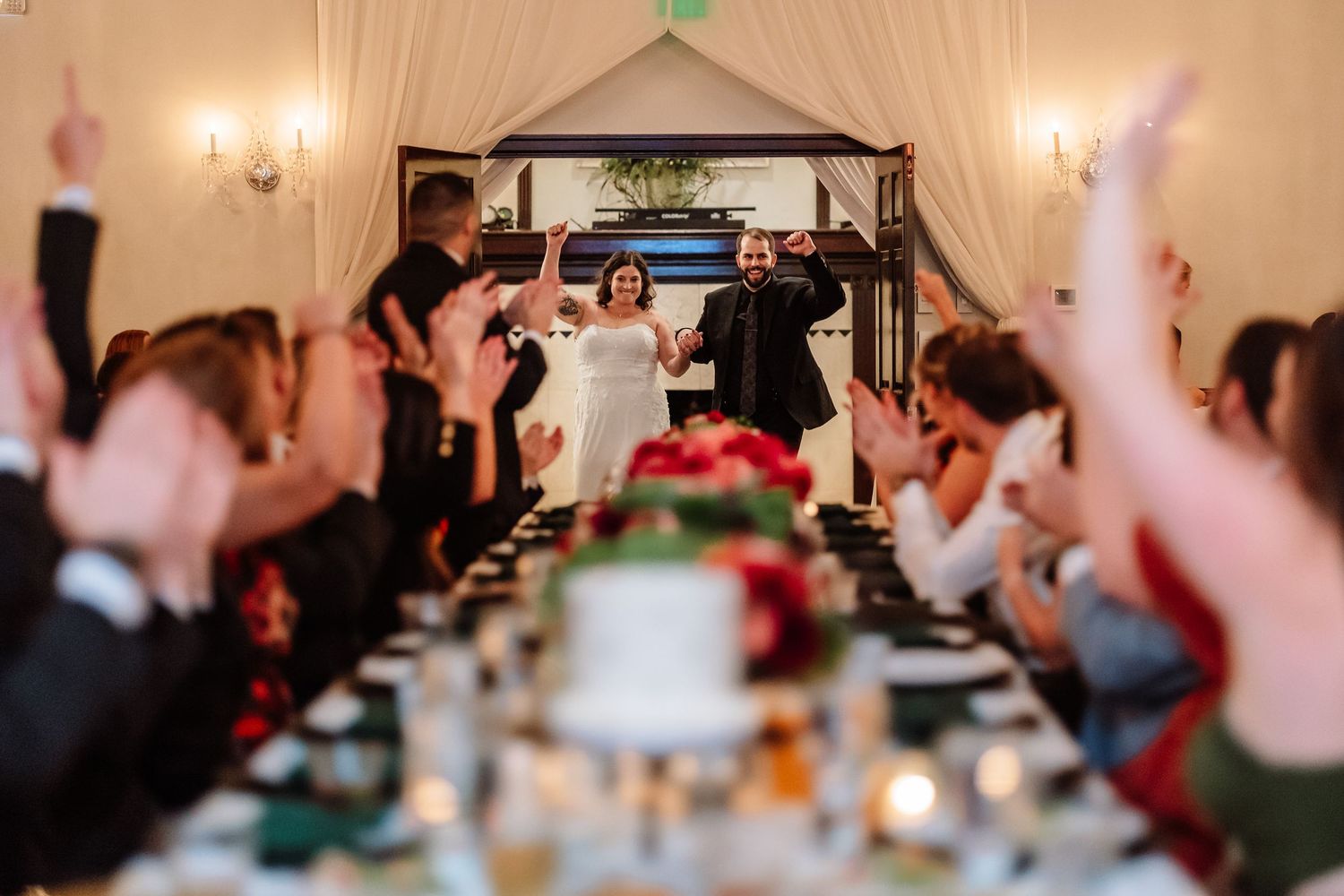 Bride and groom make grand entrance into wedding reception as guests cheer with hands raised.