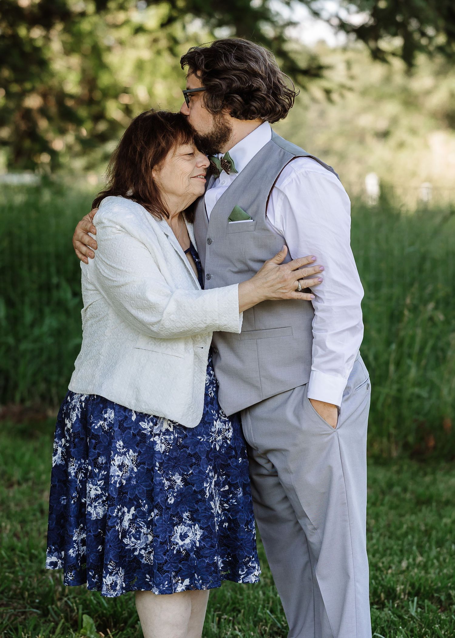 Groom in gray vest shares tender embrace with older woman in navy floral dress outdoors.