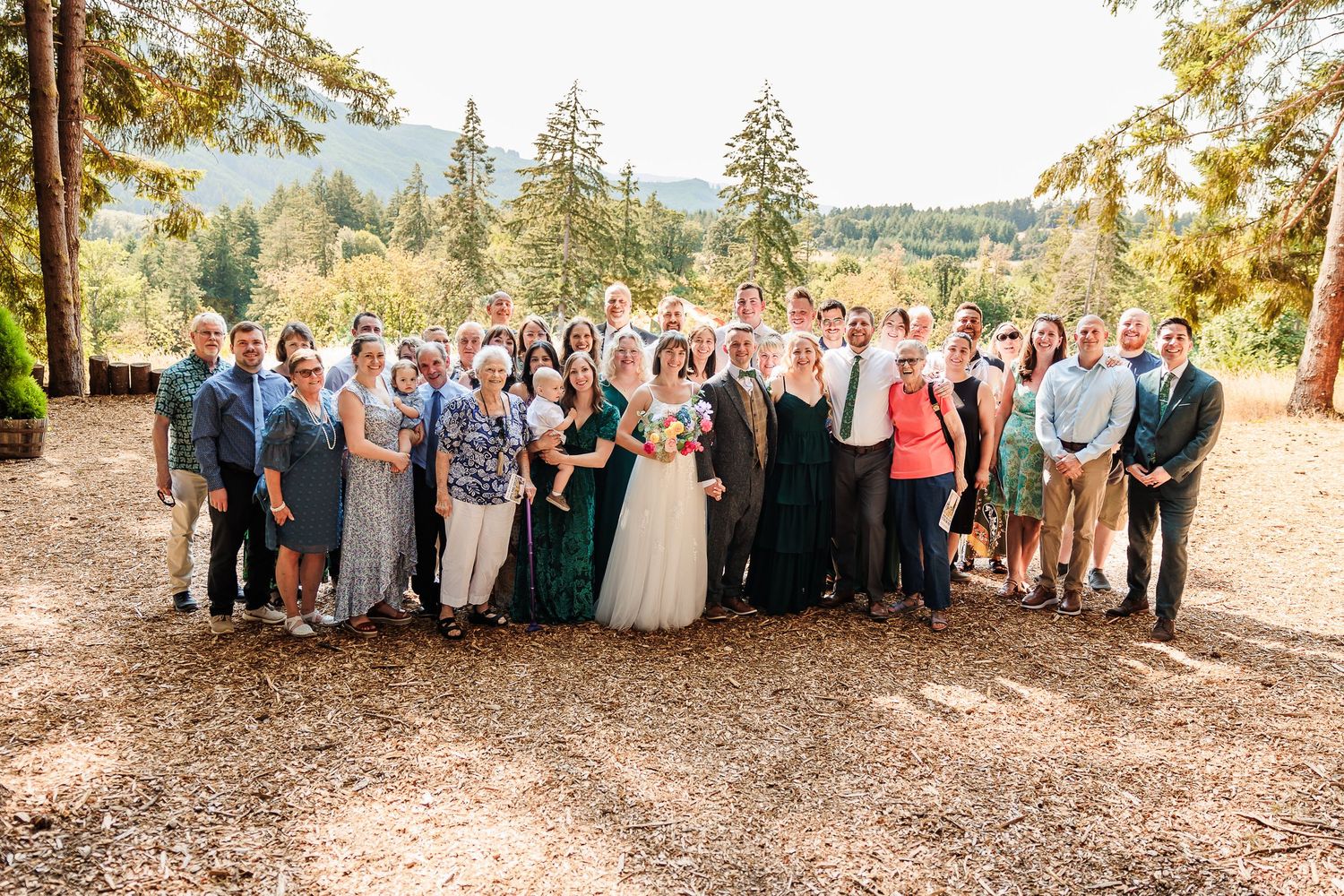 Large wedding group portrait with guests gathered outdoors in forest with mountain and tree backdrop.