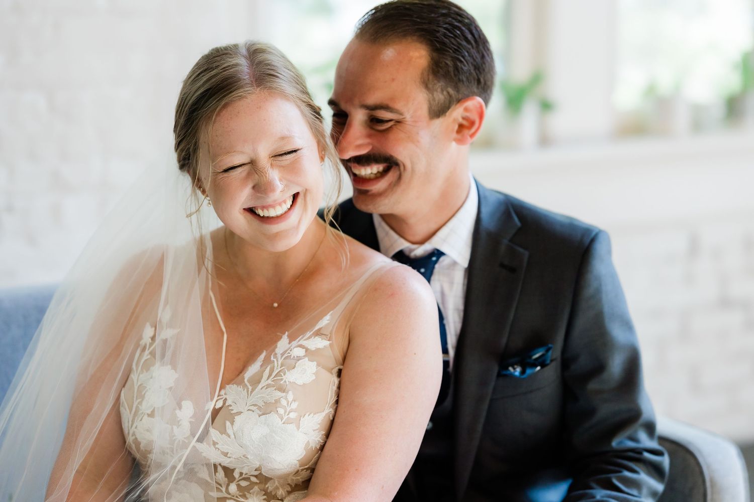 Laughing bride in lace wedding dress and veil sits close with groom in dark suit on couch.