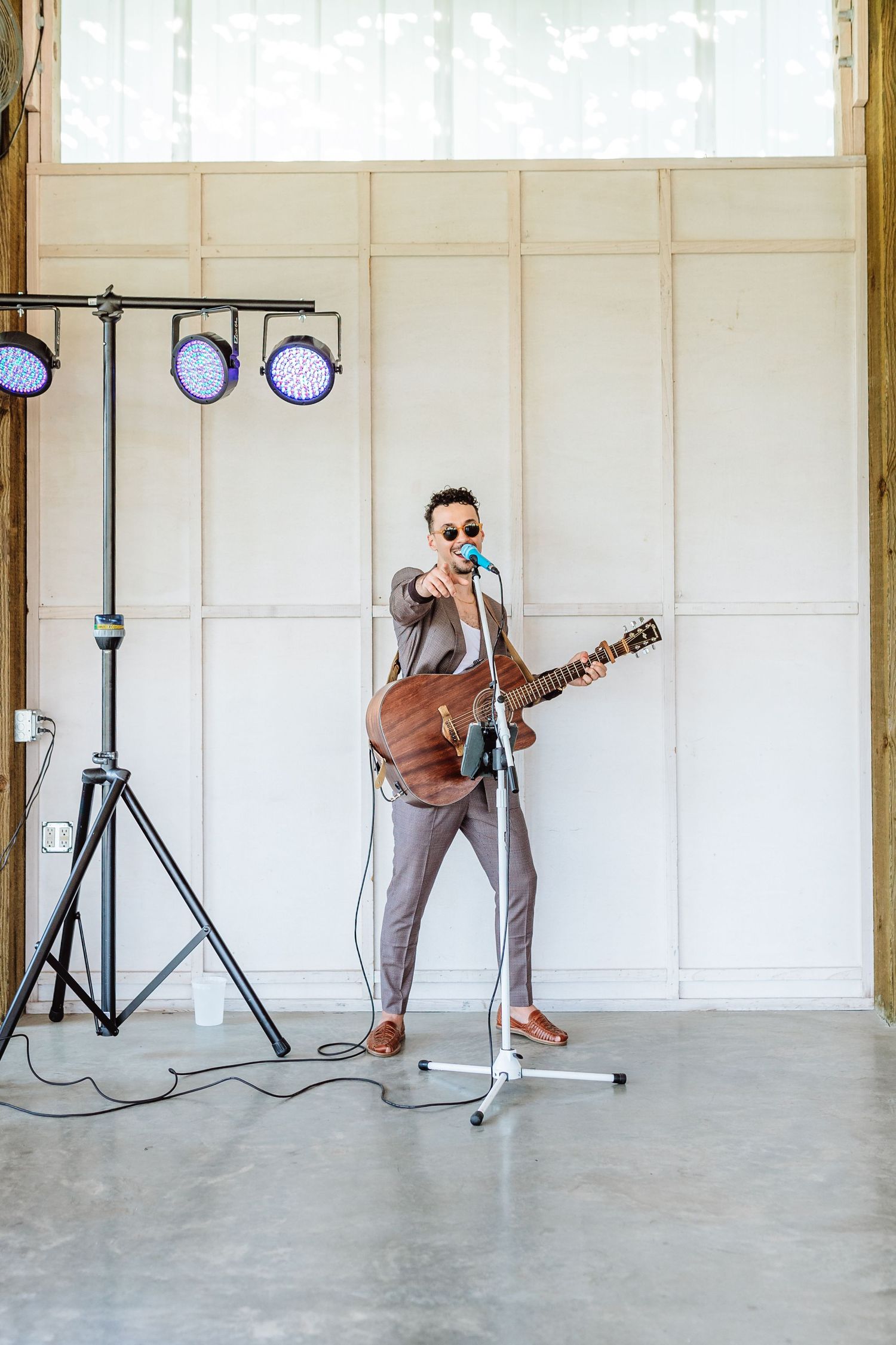 A musician in a gray suit and sunglasses plays acoustic guitar while singing into a microphone on a stand indoors.