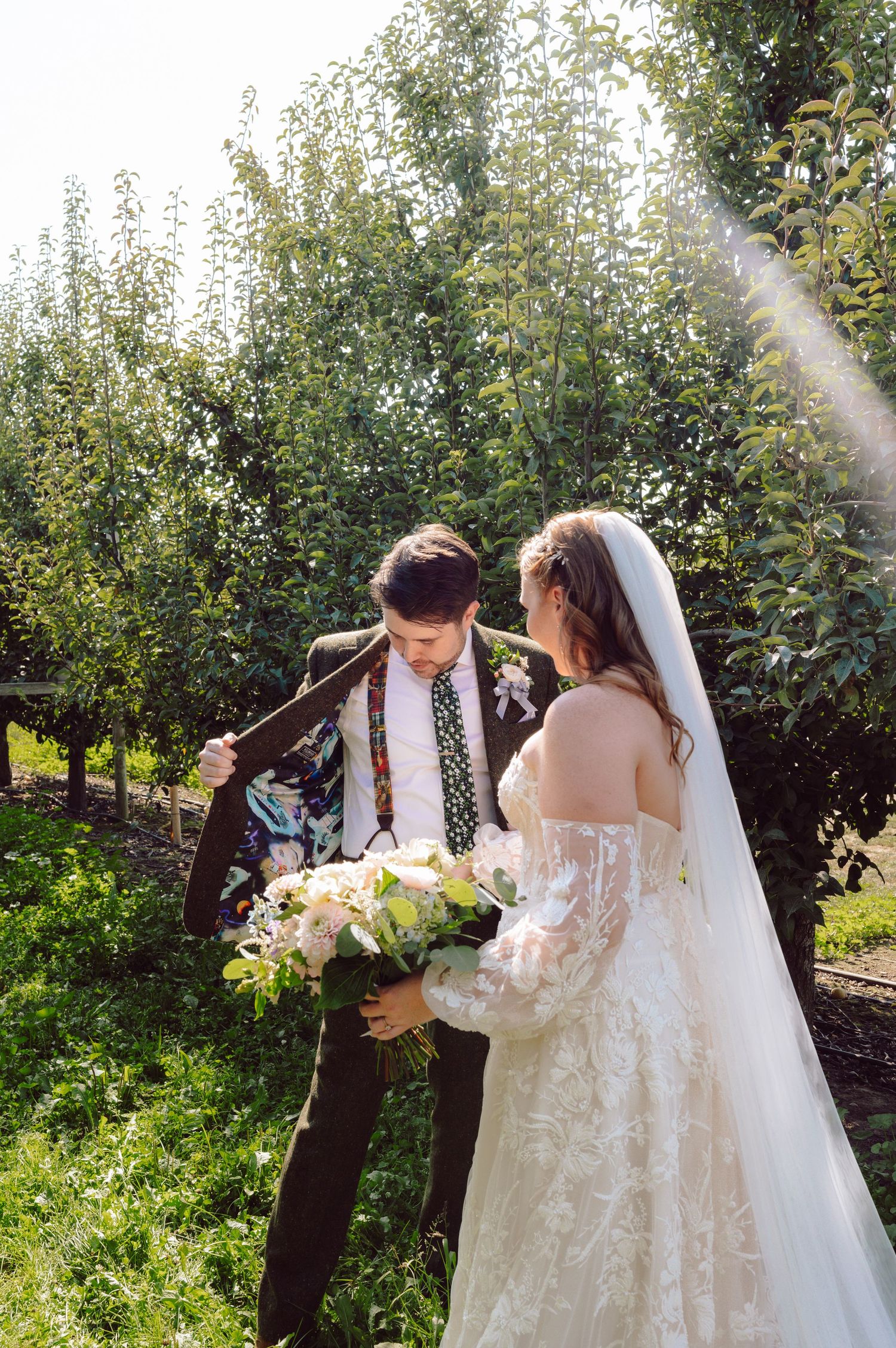 Bride in lace gown adjusts grooms floral tie in a sunlit green orchard setting.