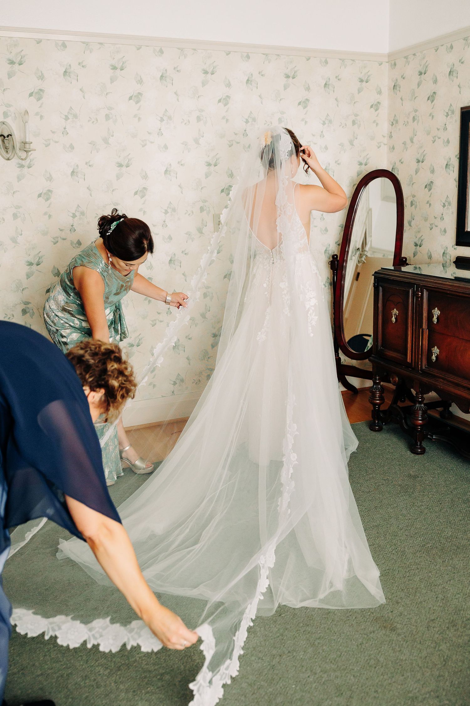 Bridesmaids helping bride arrange long lace-trimmed cathedral veil in floral wallpapered room.
