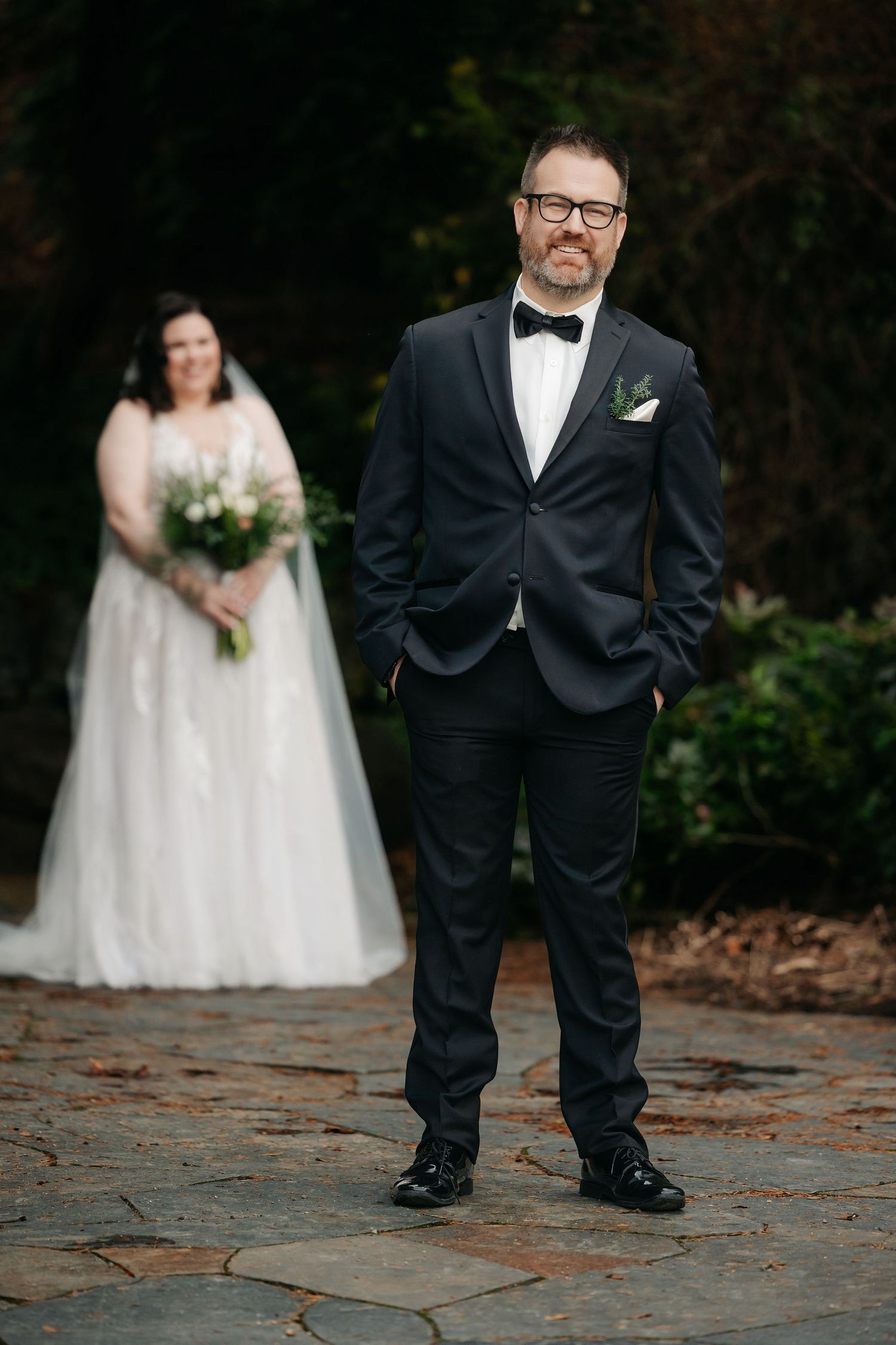 Groom in black tuxedo with bow tie stands in foreground while bride holds bouquet in soft focus behind.