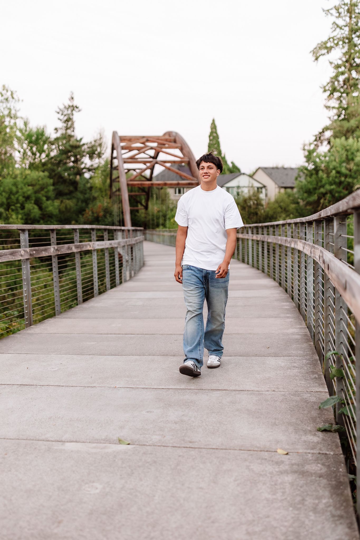 A smiling young man in a white t-shirt and jeans walks along a modern pedestrian bridge surrounded by green trees.