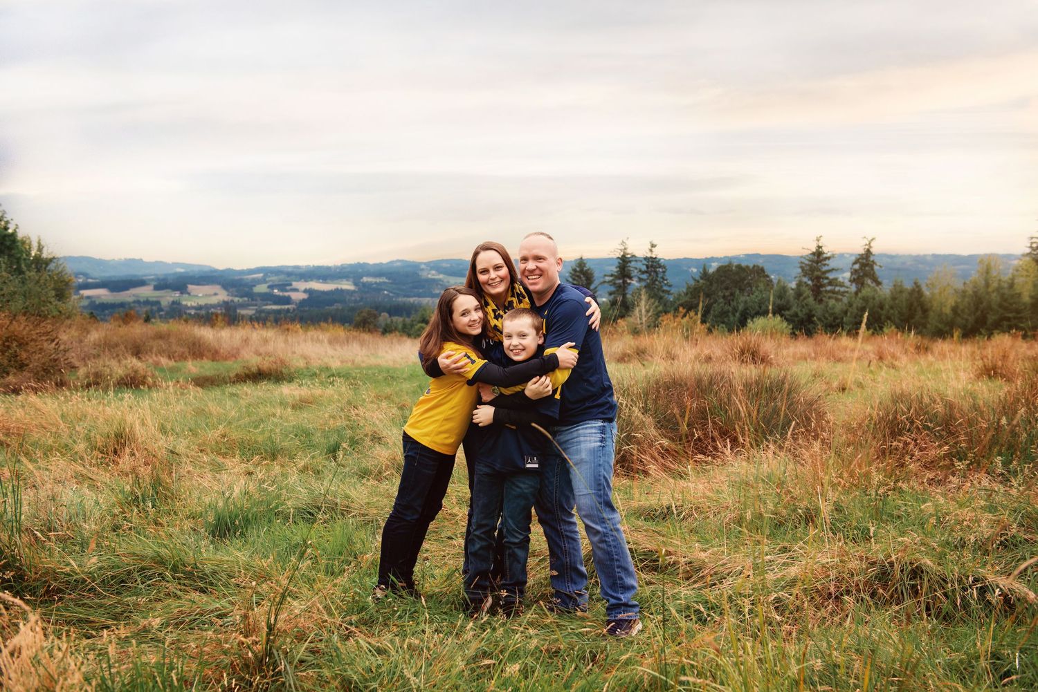 Family of four hugging and smiling in an open grassy field with scenic rolling hills at sunset.