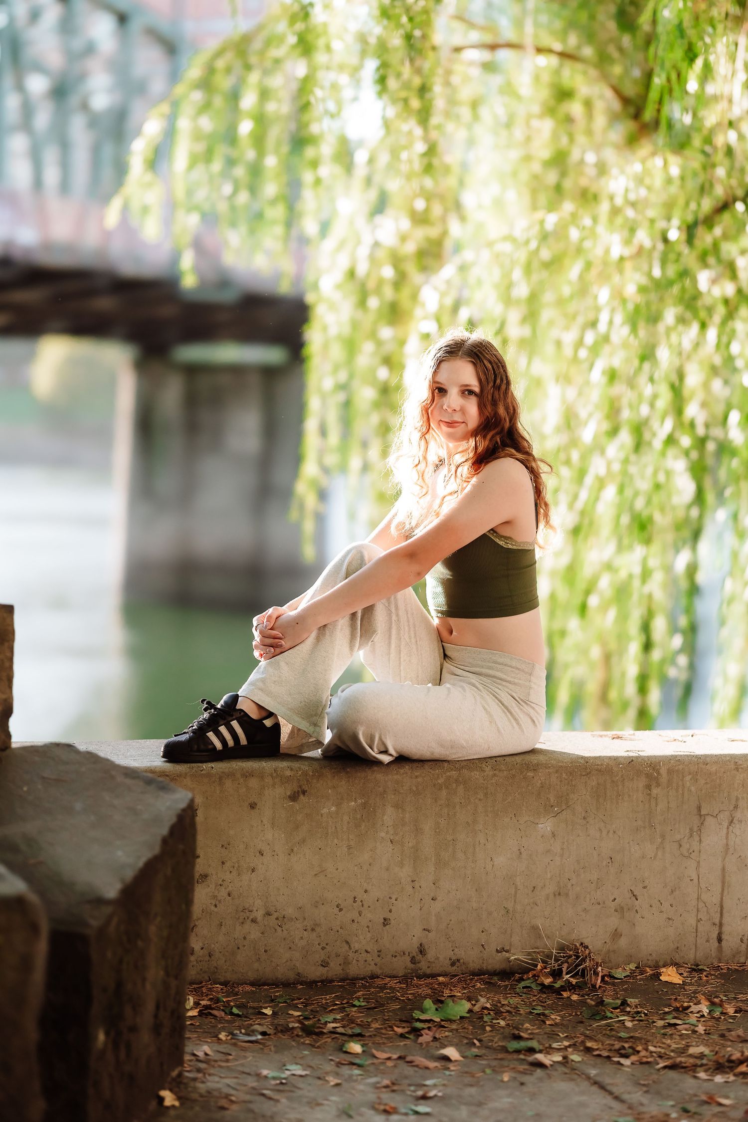 Young woman in green crop top and white pants sitting on a concrete ledge by a river under a willow tree.