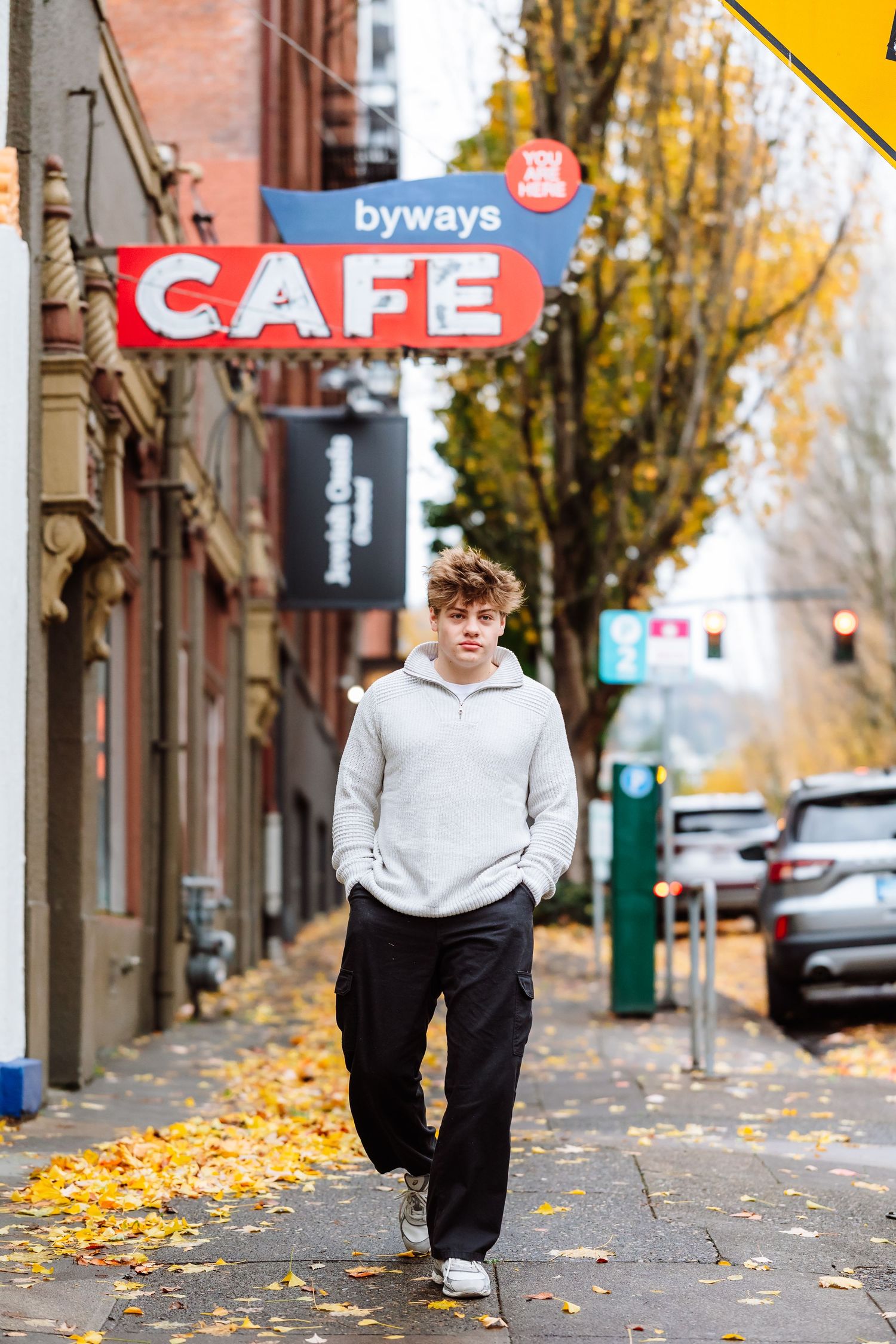 Young man in gray hoodie walking on autumn sidewalk past Byways Cafe sign in urban setting.