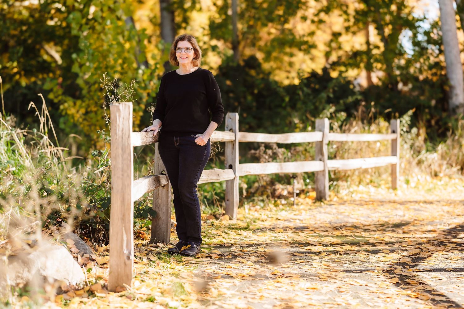 Woman in black outfit leaning on white wooden fence surrounded by golden autumn foliage on a sunlit path.