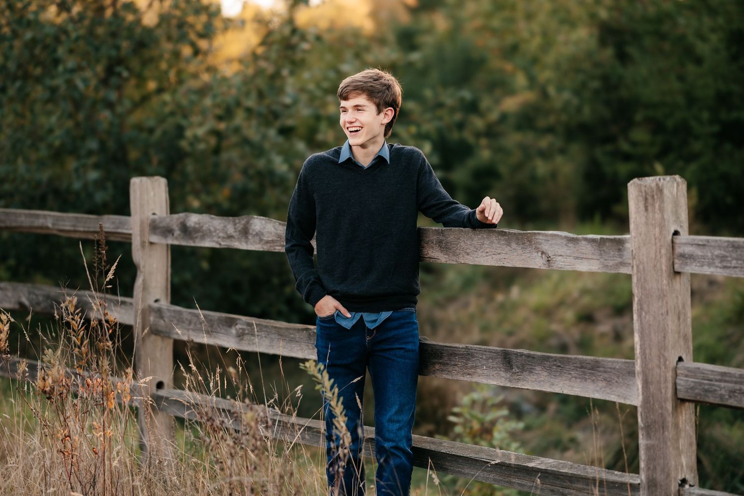 A smiling teenage boy in a black sweater leans against a rustic wooden fence outdoors in a natural setting.