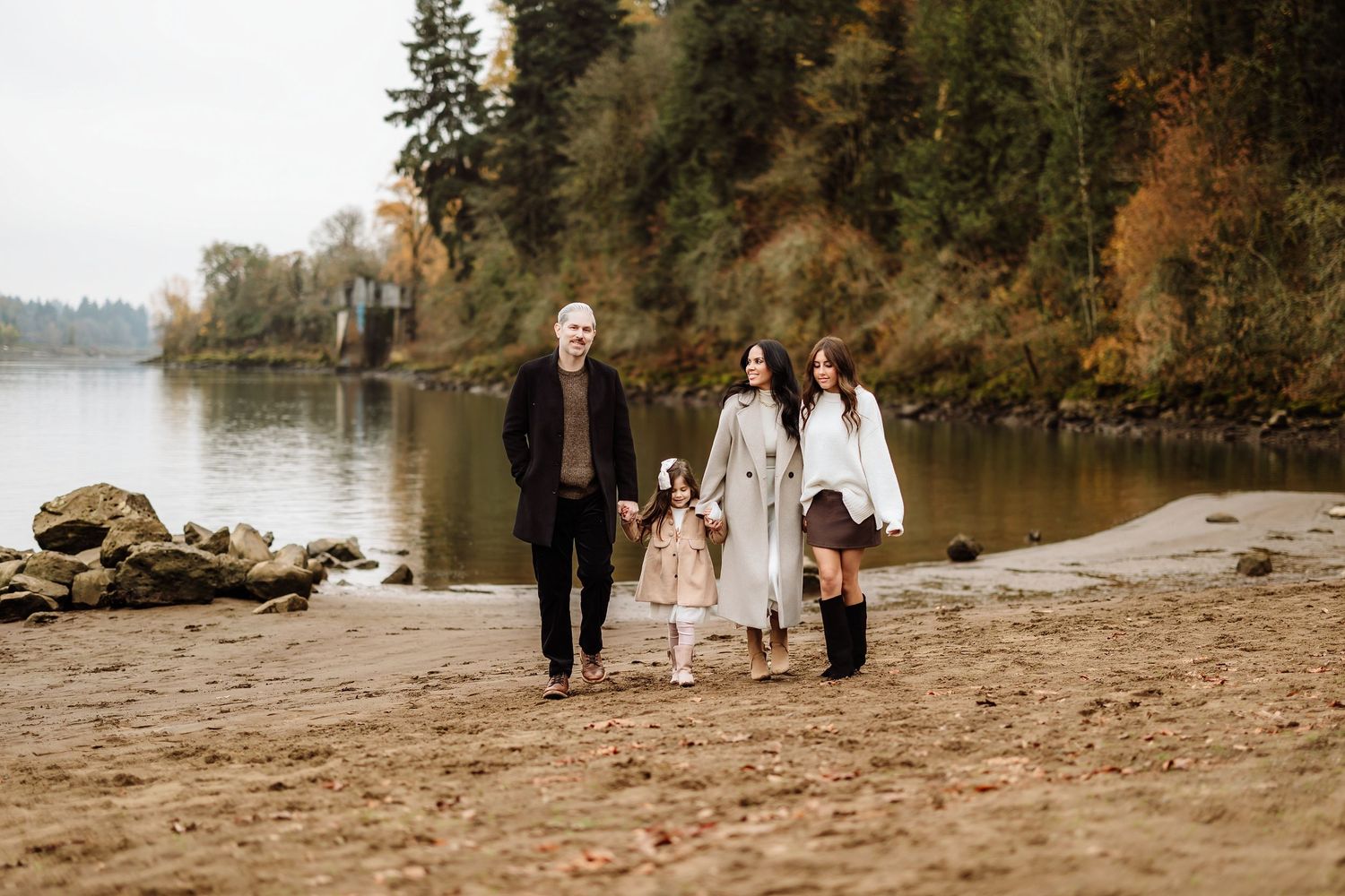 Family of three walking along a sandy riverbank surrounded by autumn foliage on an overcast day.