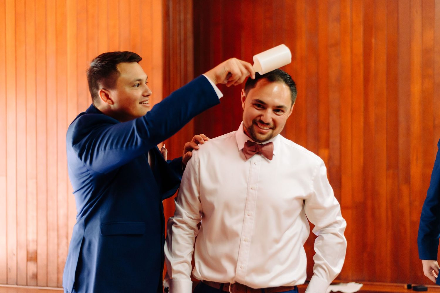 Groomsman using a lint roller on groom in blue suit and bow tie before wedding ceremony.