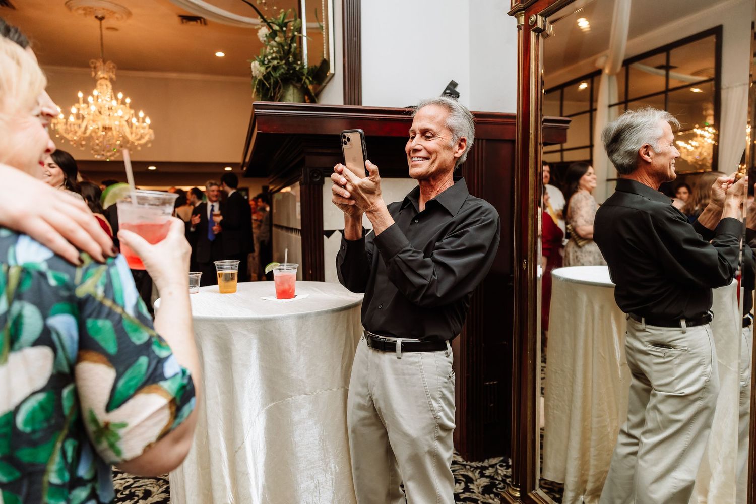 Smiling man in black shirt taking photos on smartphone at an elegant wedding reception cocktail hour.