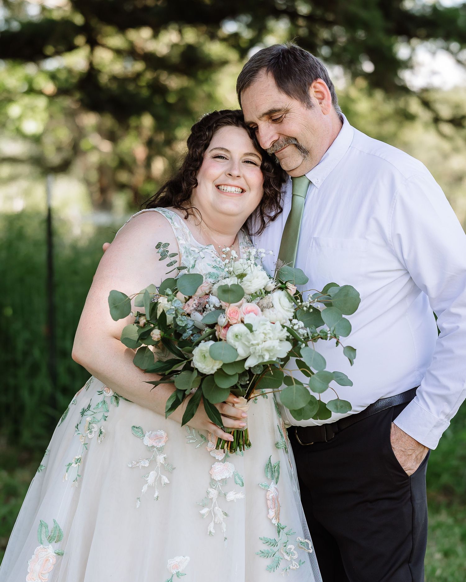 Smiling bride in floral gown holds bouquet as groom in green tie embraces her outdoors.