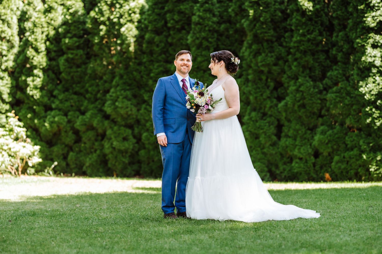 Bride holding floral bouquet and groom in blue suit share a tender moment on a lush green lawn.