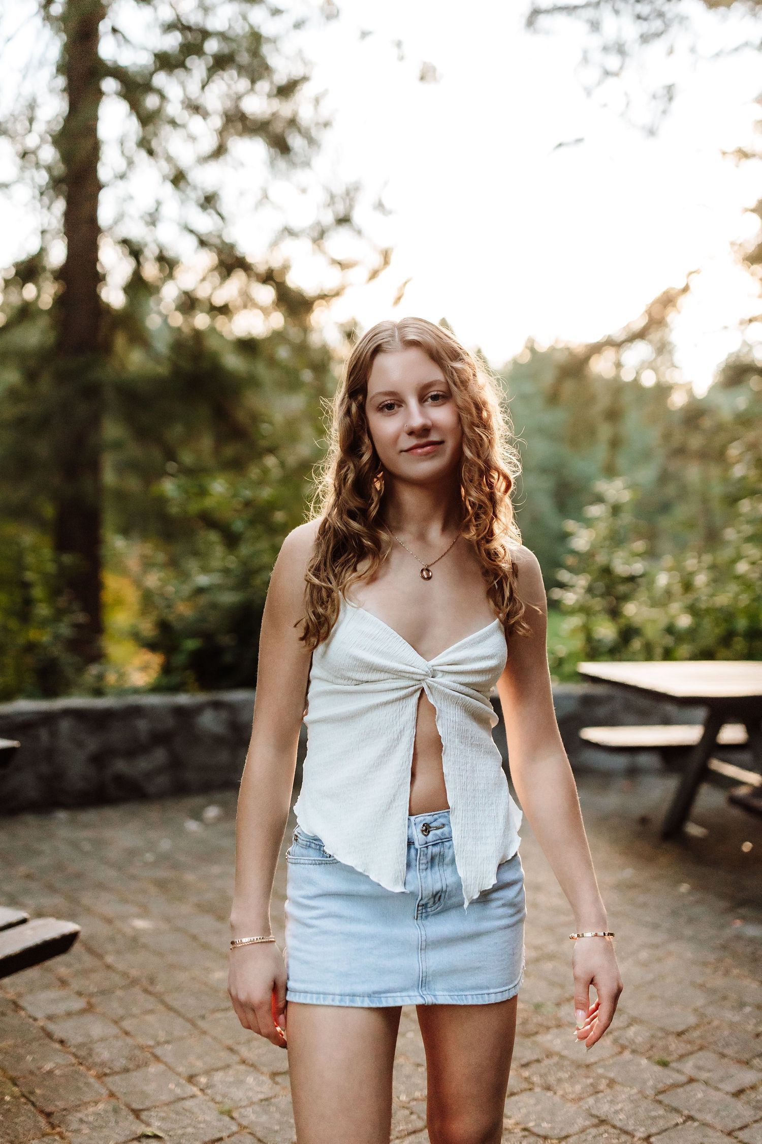Young woman with curly hair wearing a white crop top and denim mini skirt standing outdoors in golden hour light.