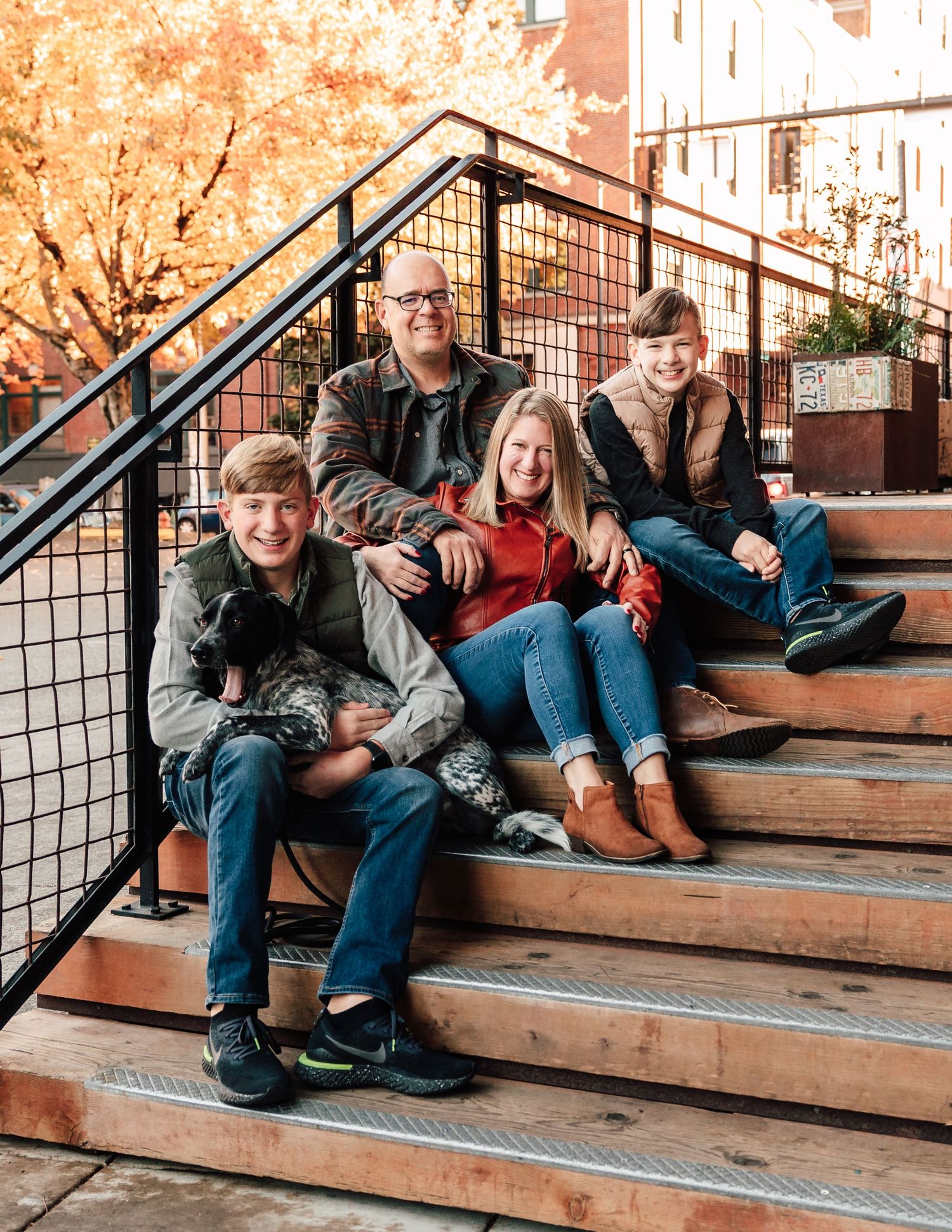 Family of four smiling on outdoor steps surrounded by autumn foliage in an urban setting.