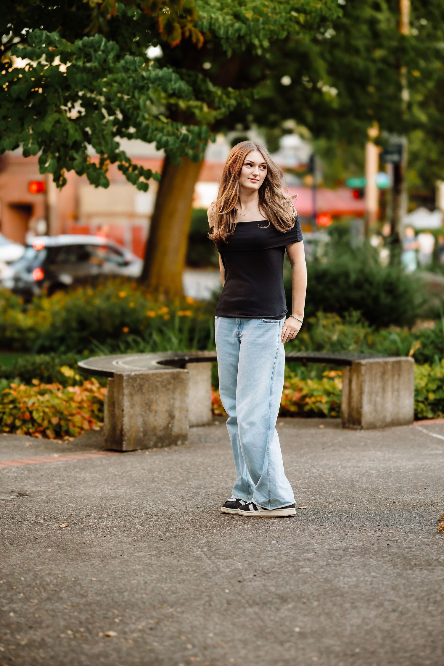 Young woman in black top and wide-leg jeans standing in an urban park with lush green trees and colorful flowers.