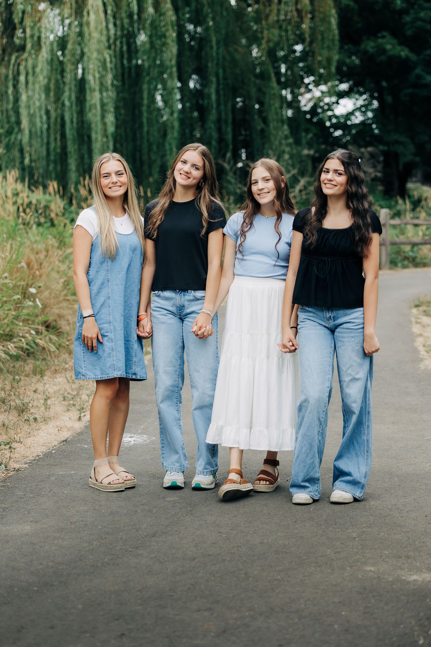 Four young women smiling and walking together on a path surrounded by lush green trees on a sunny day.