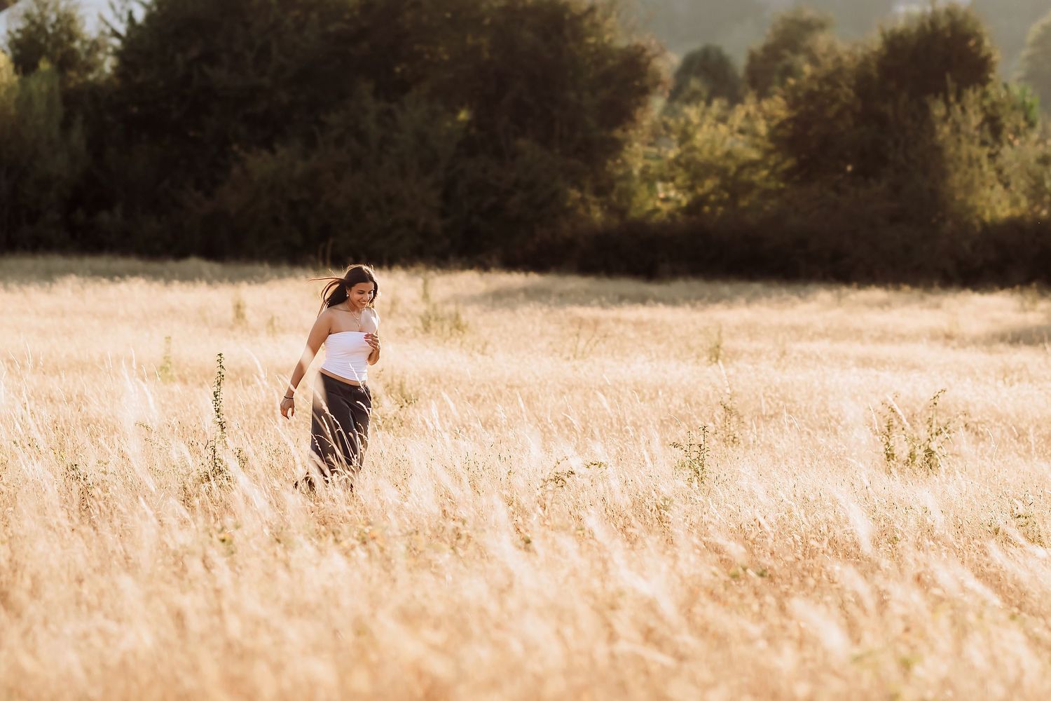 A woman in a white top and dark skirt walks through a golden wheat field with green trees in the background.