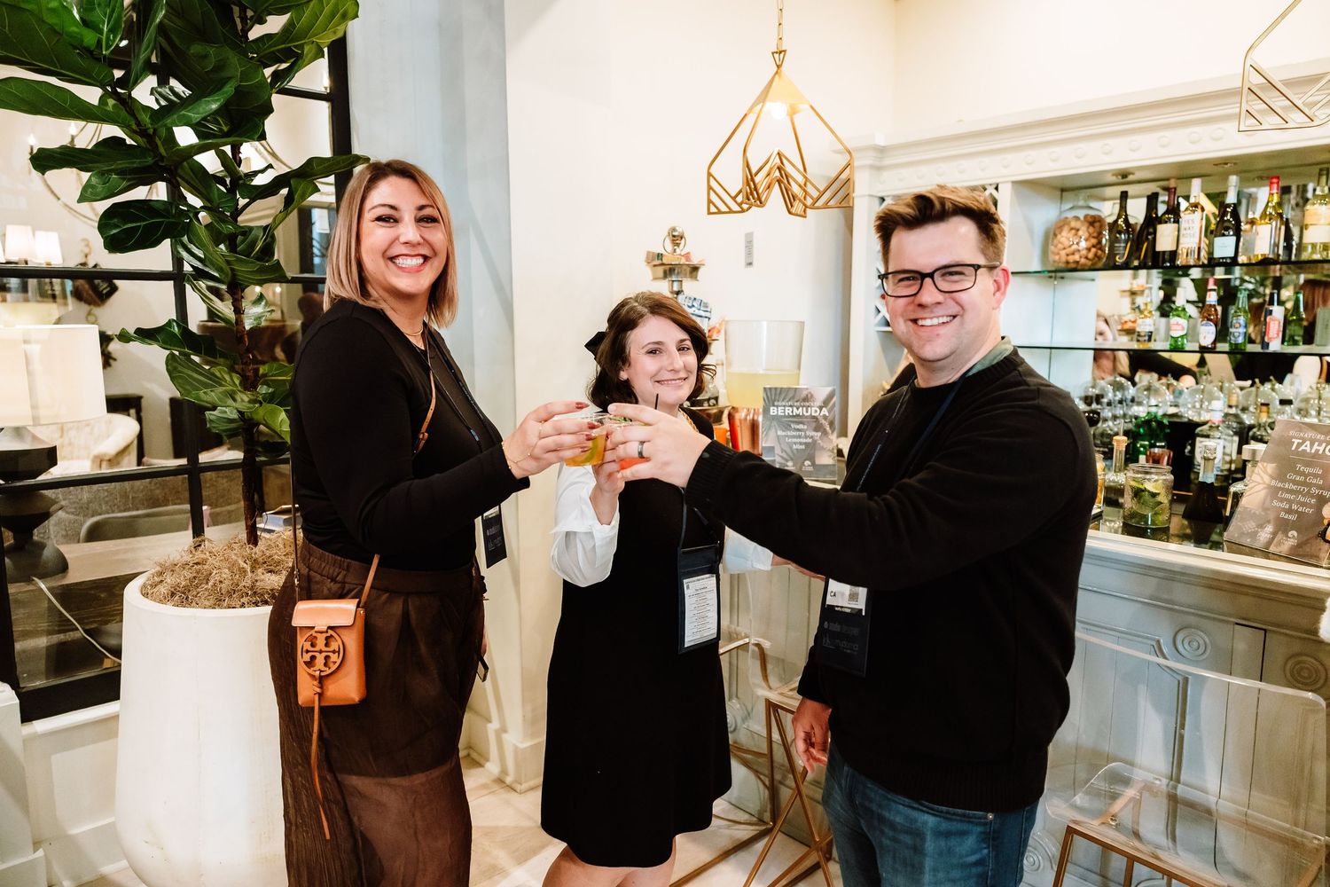 Three people smile and toast with drinks inside a stylish home decor boutique.