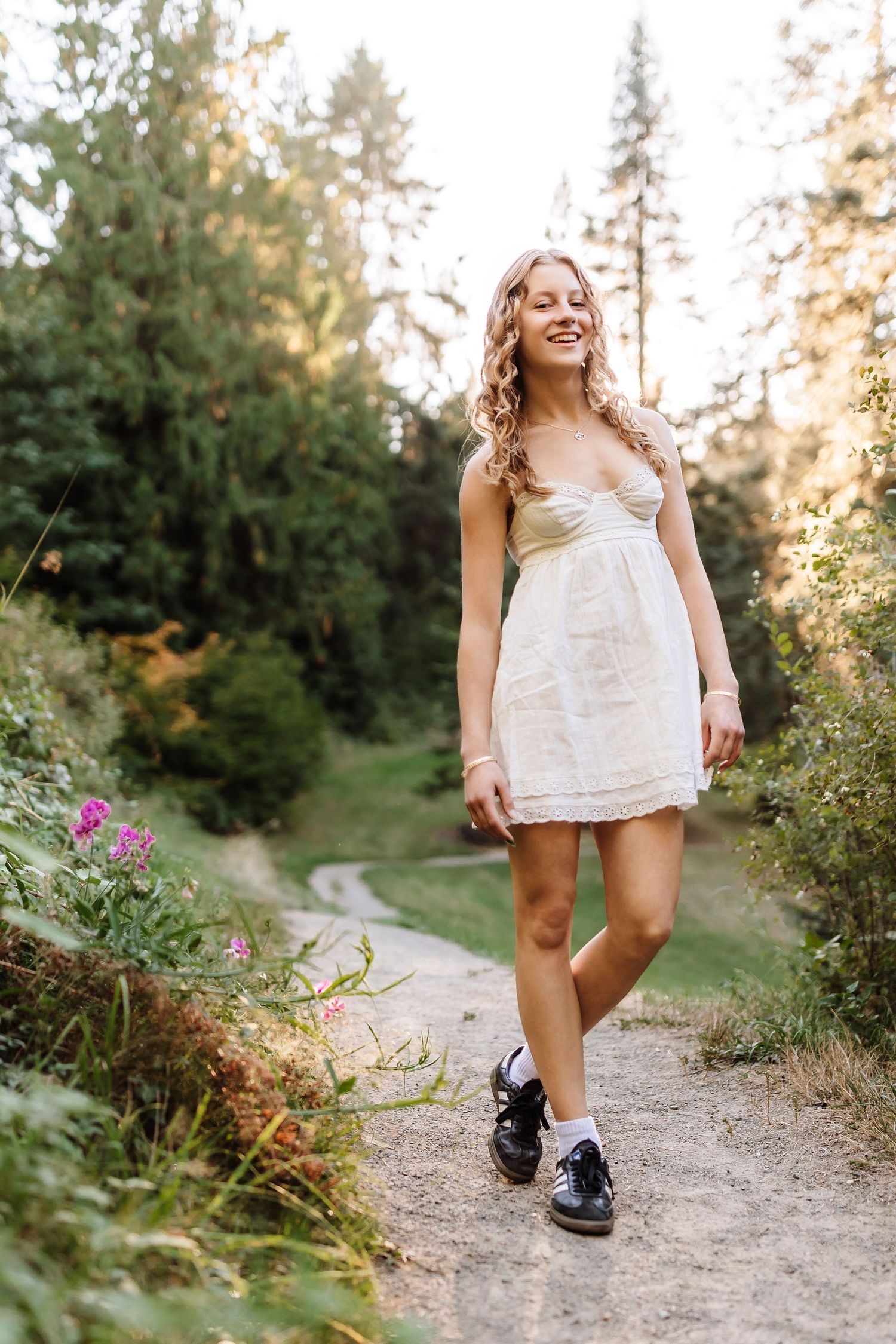 Young woman in white mini dress and black shoes smiling on a forest path surrounded by lush greenery.