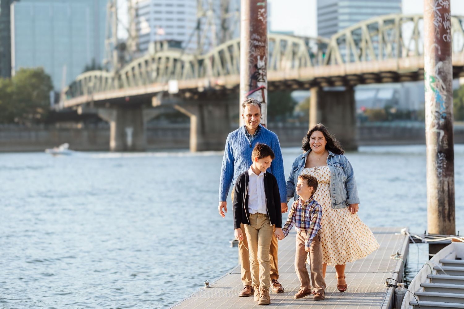 Happy family of four walking along a waterfront boardwalk with a bridge in the background.