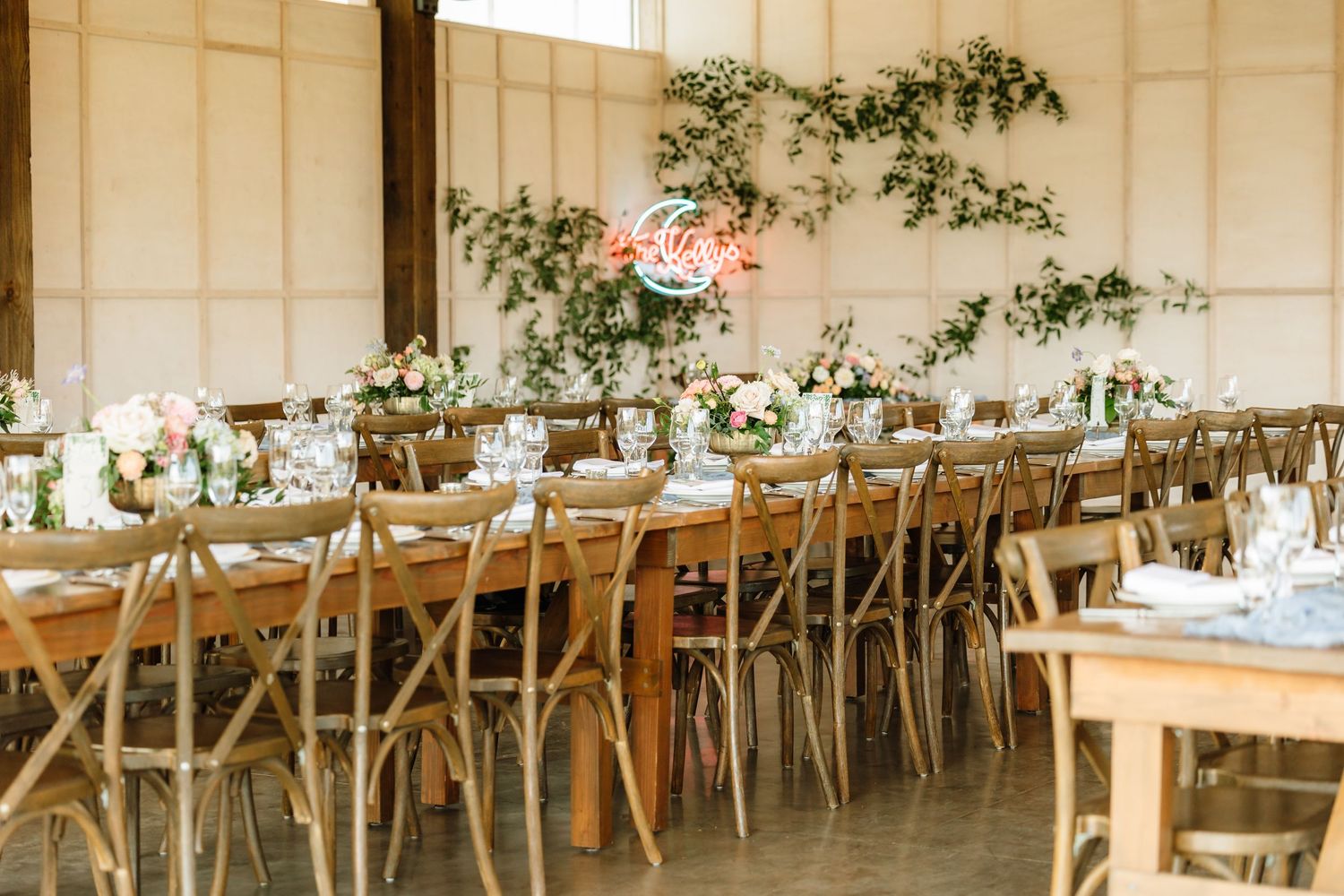 Rustic wedding reception hall with long wooden tables, cross-back chairs, floral centerpieces, and neon sign.